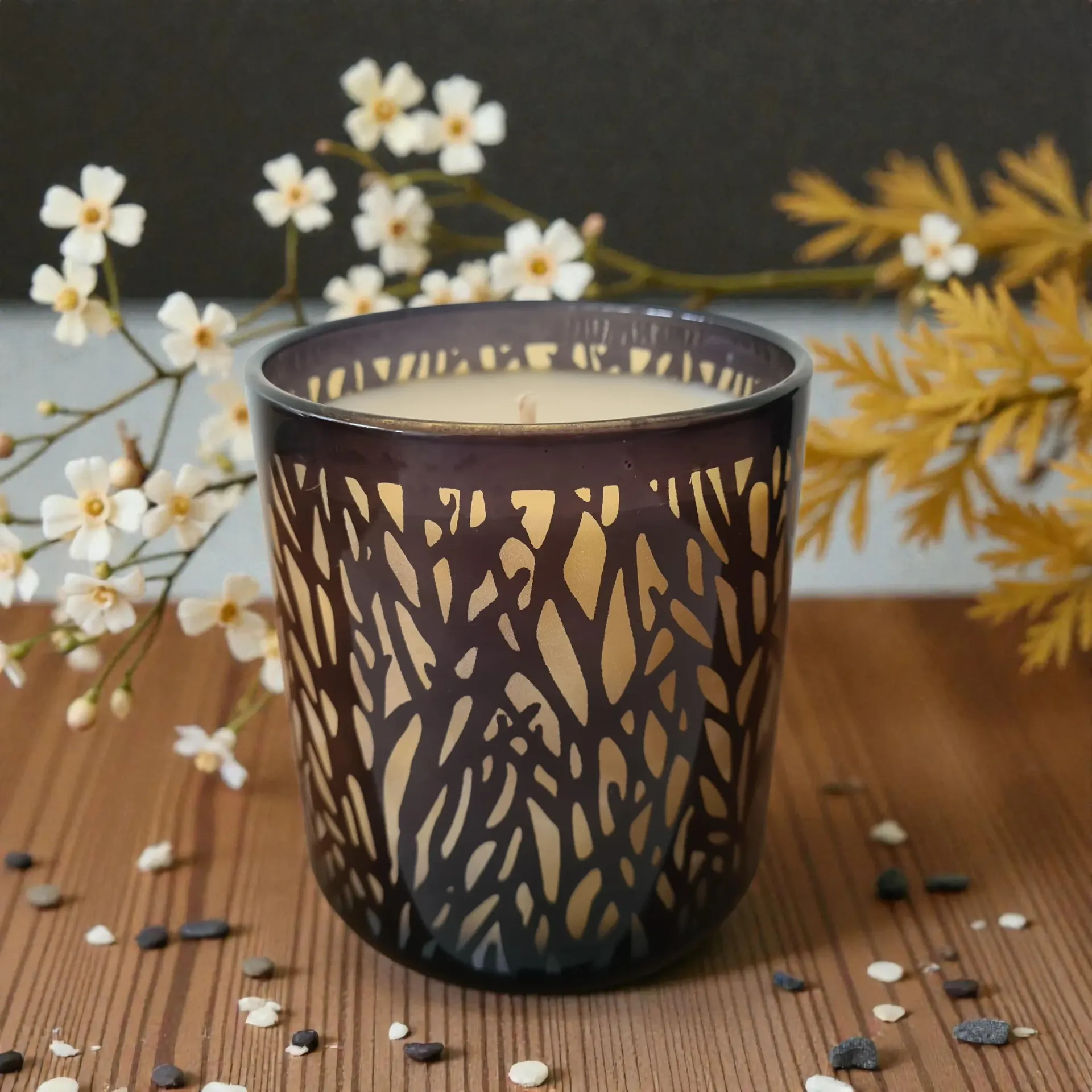 Candle in a dark, patterned glass holder, surrounded by flowers and small pebbles on a wooden surface.