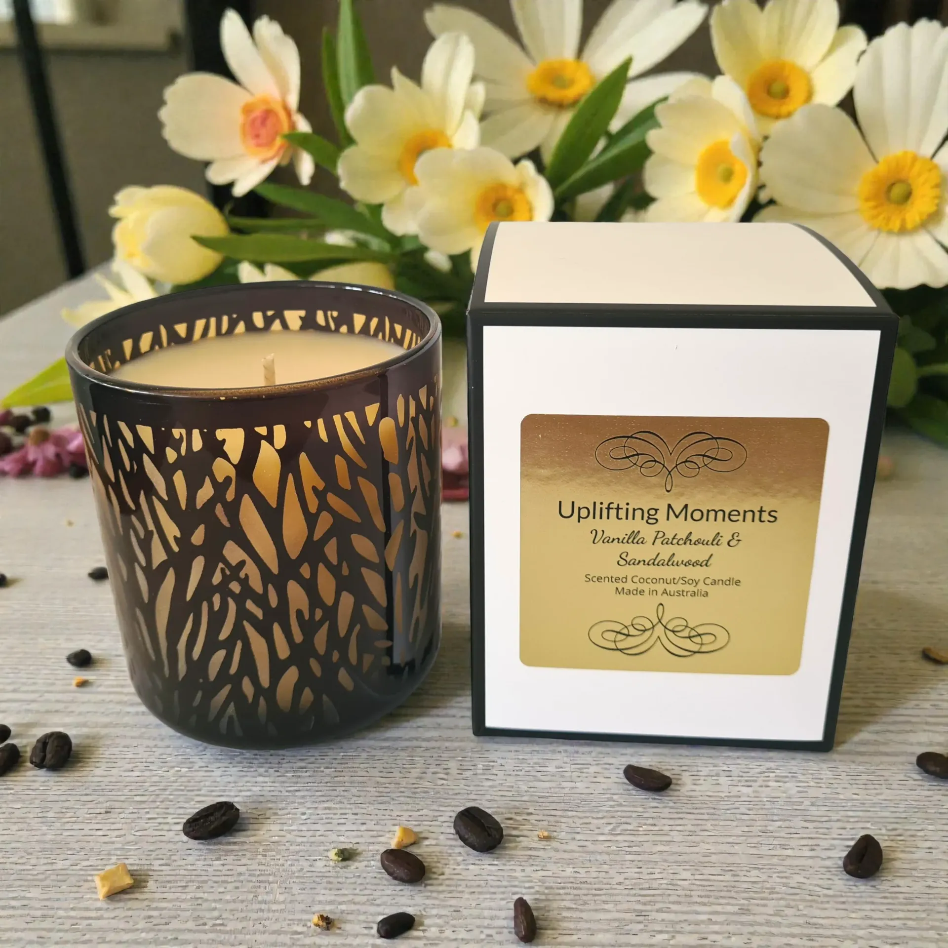 Candle in a dark, patterned glass holder, surrounded by flowers and small pebbles on a wooden surface.