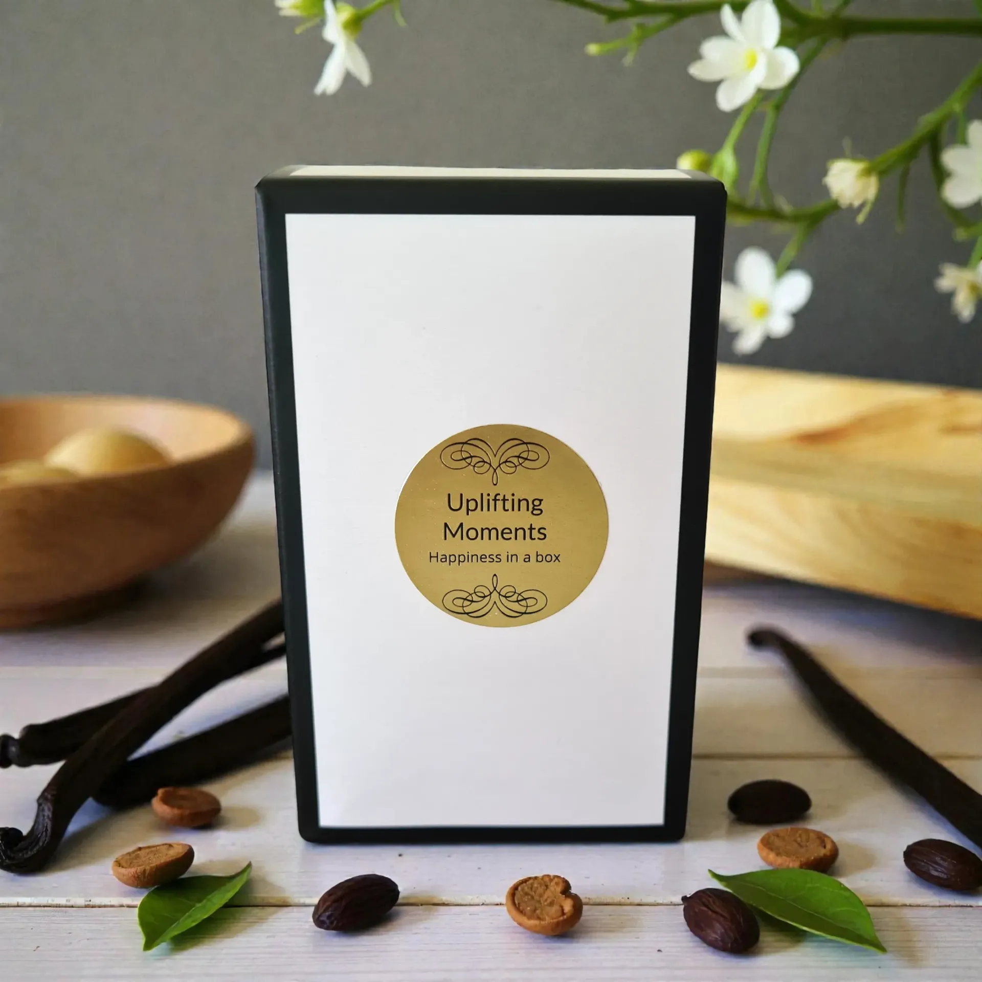 White candle with gold label, vanilla beans, and flowers on wooden surface.
