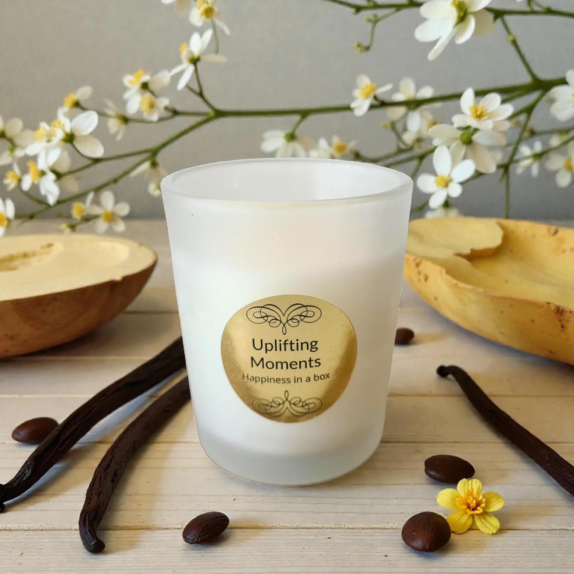 White candle with gold label, vanilla beans, and flowers on wooden surface.