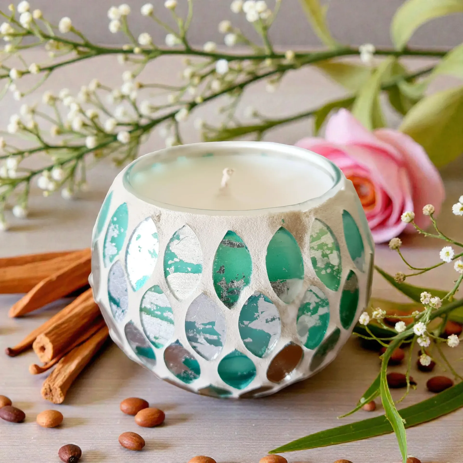 A candle in a teal mosaic glass container, with cinnamon sticks, flowers, and seeds.