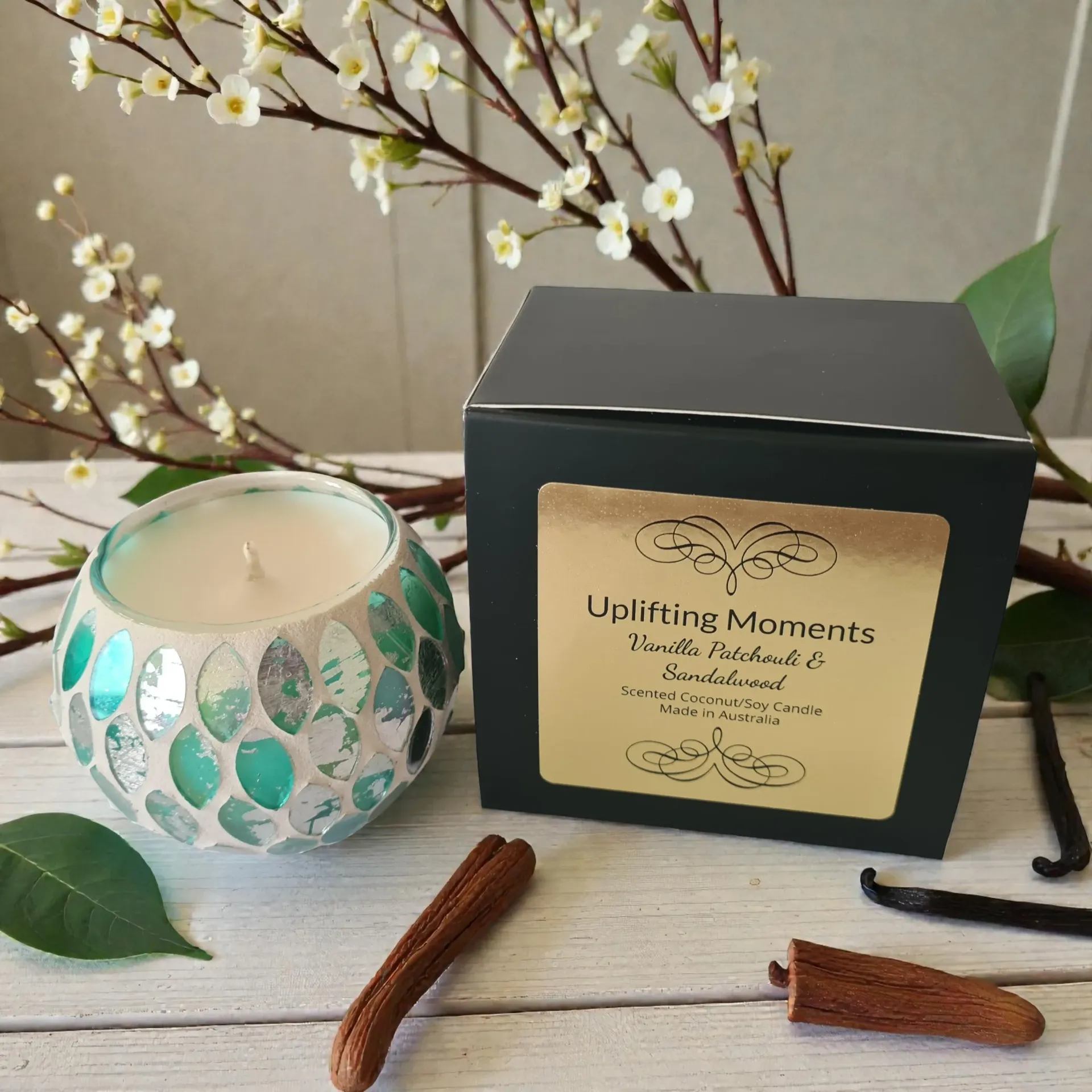 A candle in a teal mosaic glass container, with cinnamon sticks, flowers, and seeds.