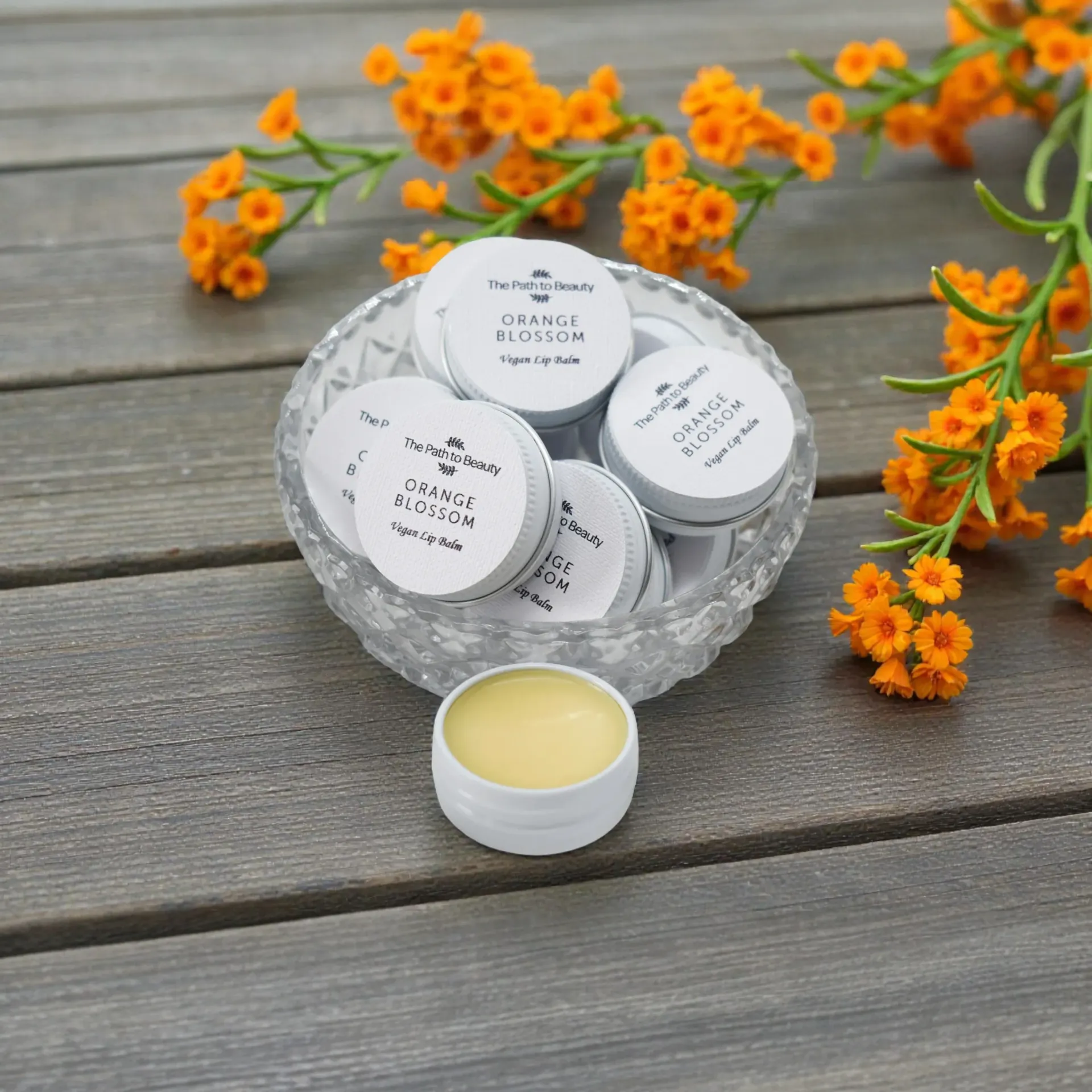 White lip balm tins in a glass bowl on wood with orange flowers.