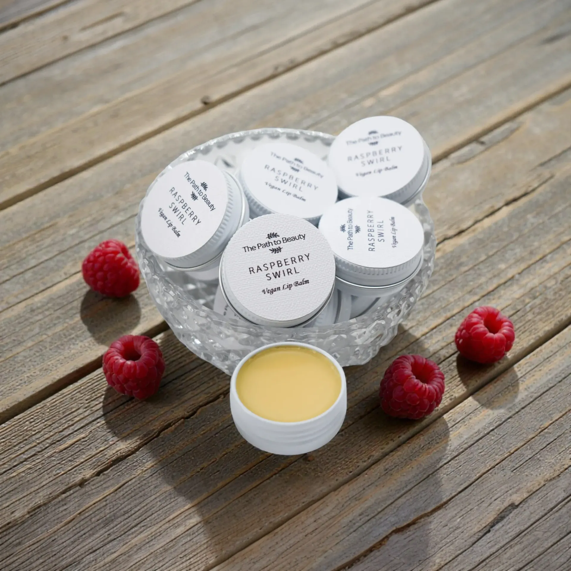 Lip balms in small tins, with raspberries, on a wooden surface.