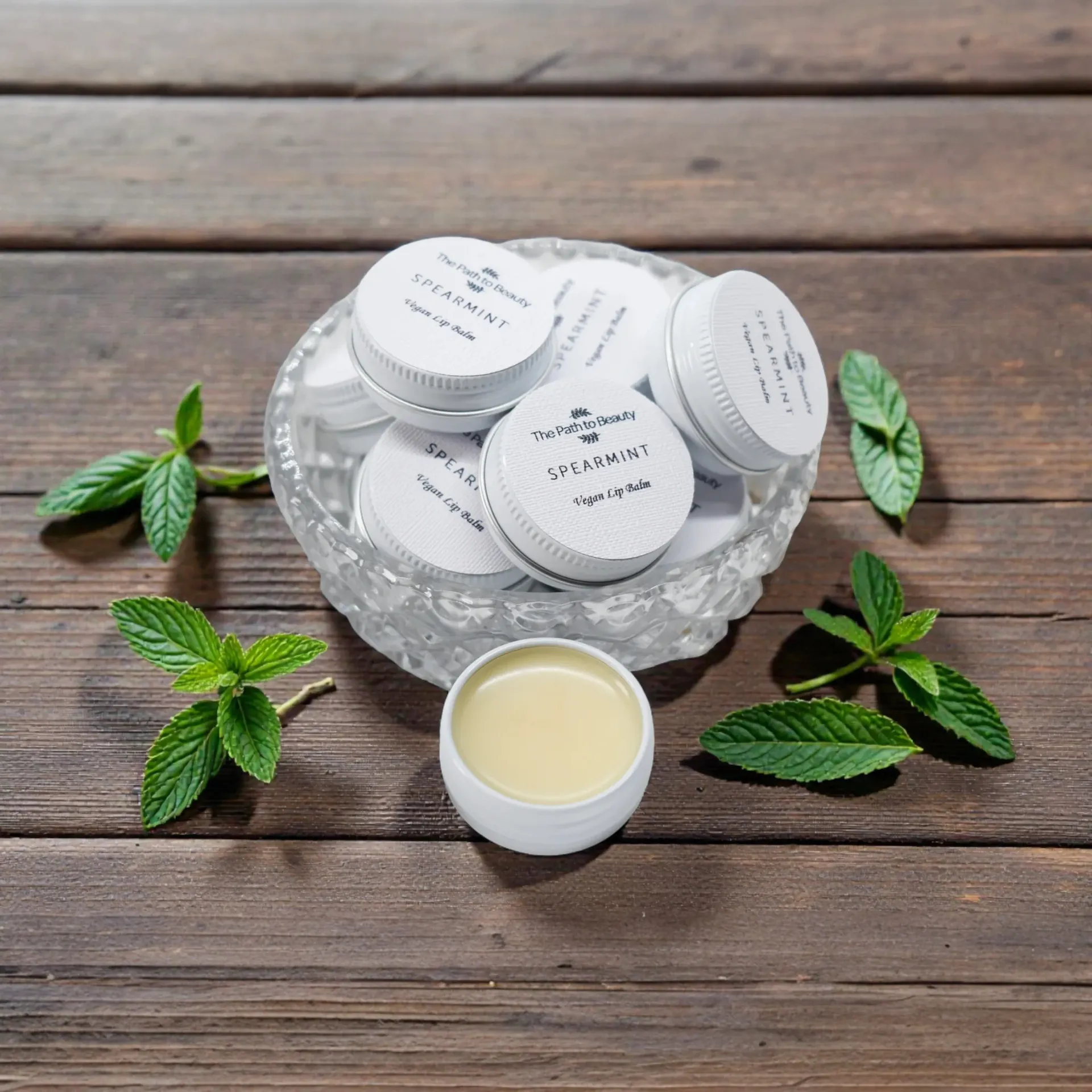 Small white jars of balm in a glass bowl, surrounded by fresh mint sprigs on a wooden surface.