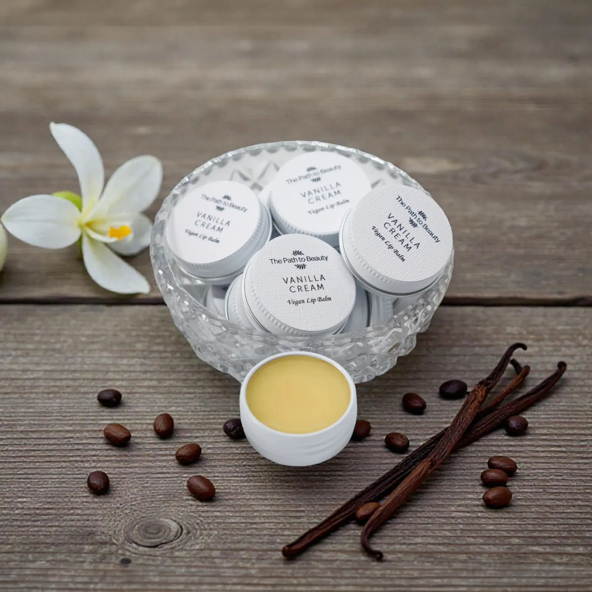 Vanilla cream jars in a crystal bowl, with vanilla beans, coffee beans, and flower on a wooden surface.
