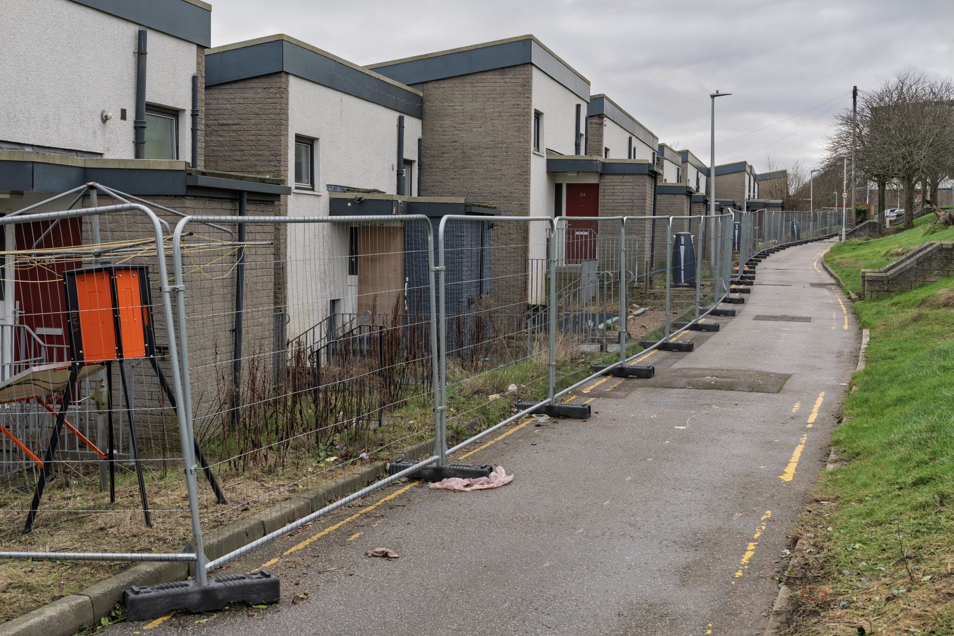 Row of townhouses behind temporary fencing along a sidewalk. Gray and white buildings, overcast sky.
