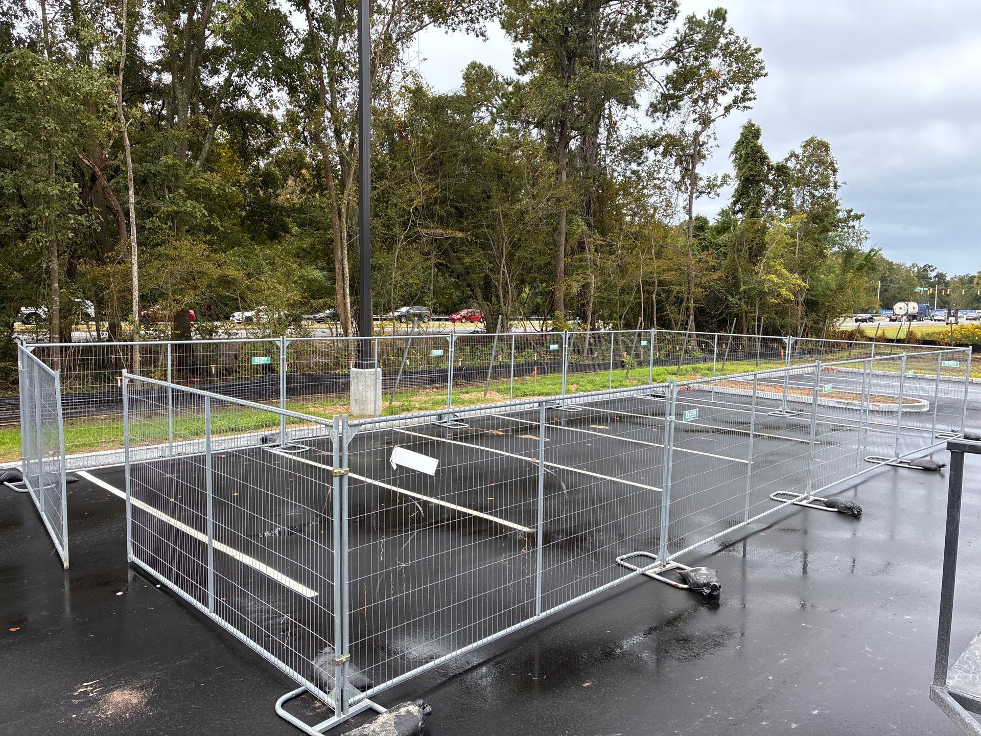 Metal temporary fencing surrounds an empty asphalt area, possibly a parking lot. Trees in the background.
