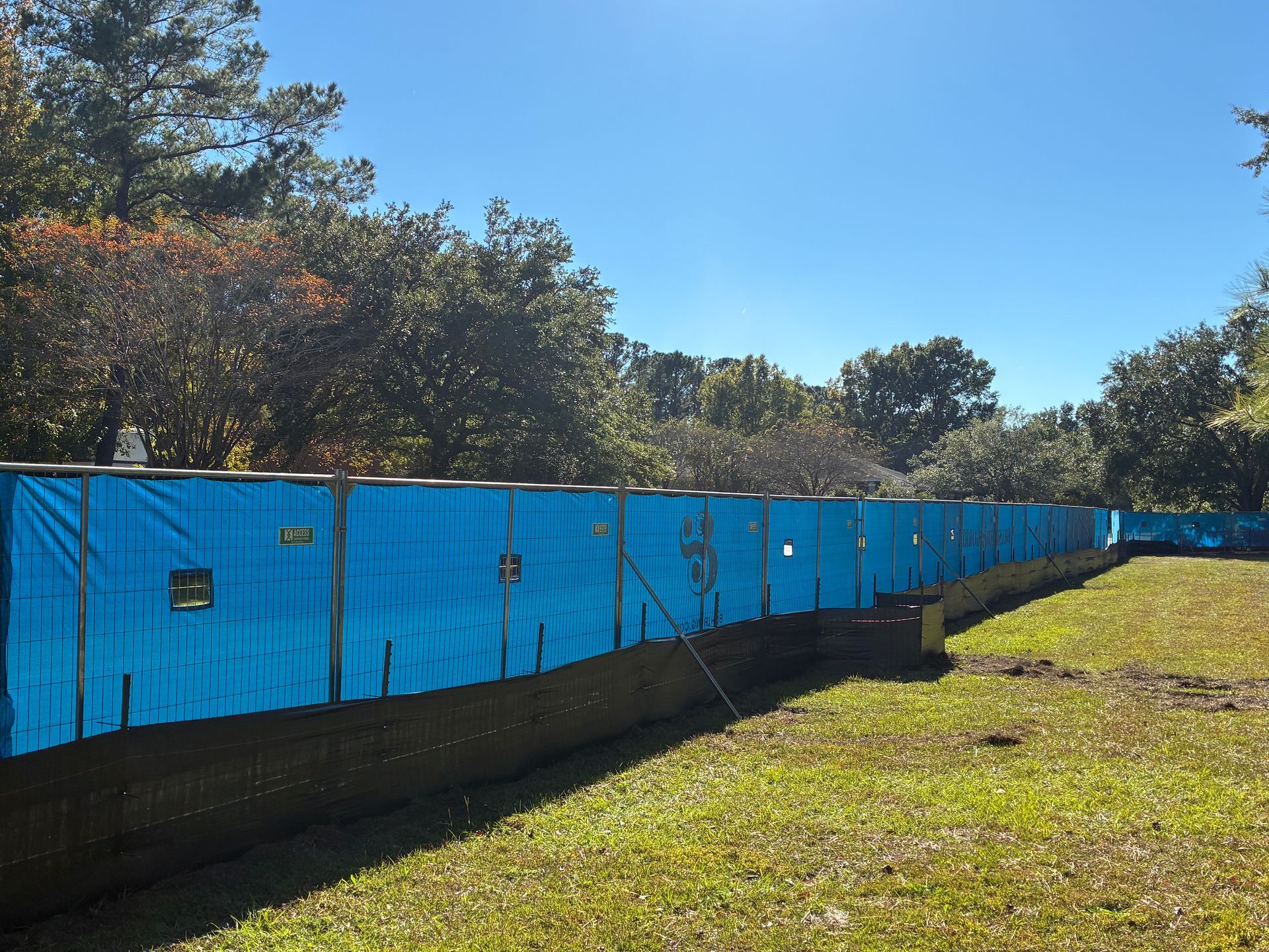 Blue tarp fencing along a grassy area, trees in the background, clear blue sky.