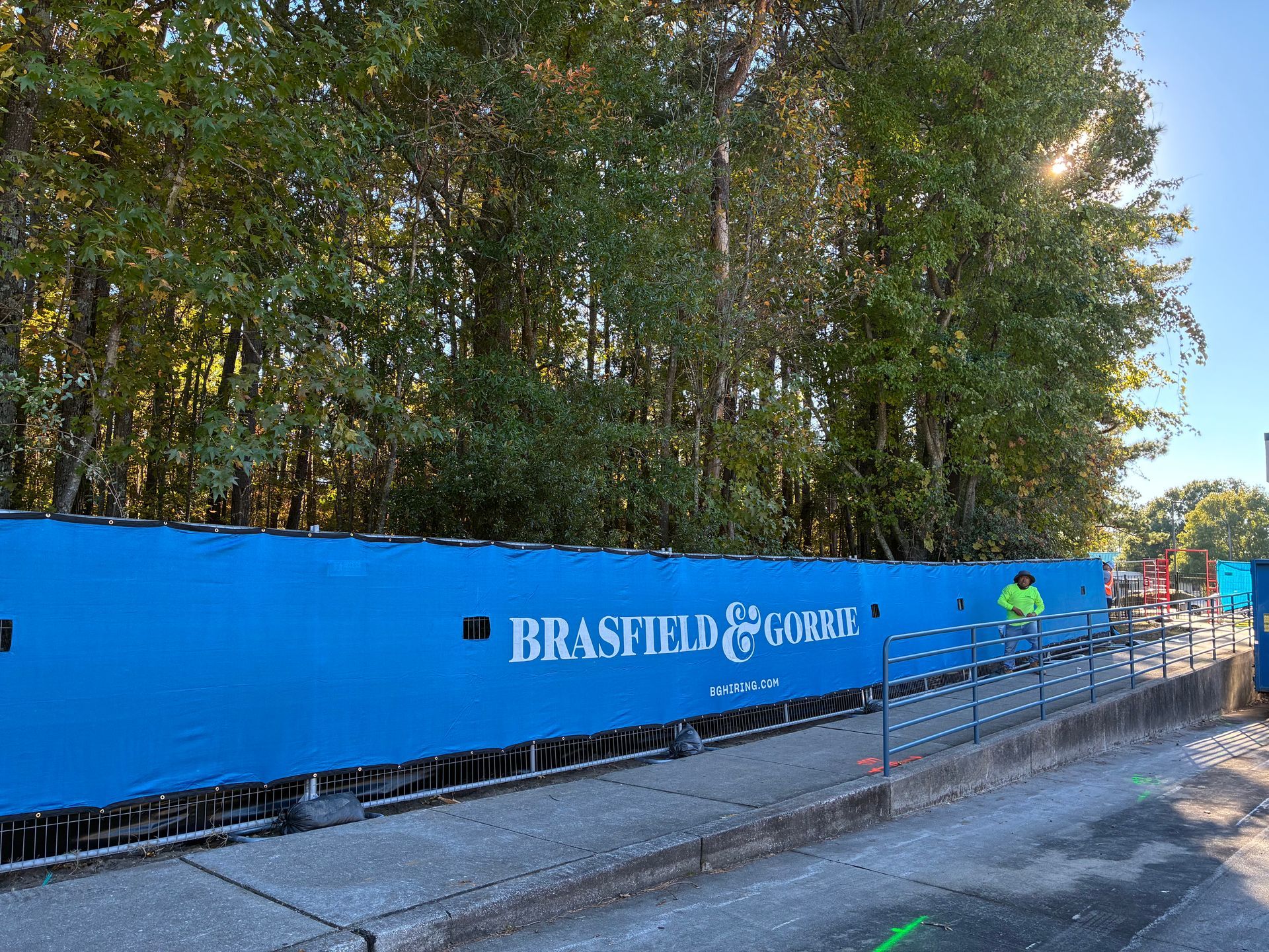 Blue construction barrier with Brasfield & Gorrie text, trees in background, person in yellow vest.