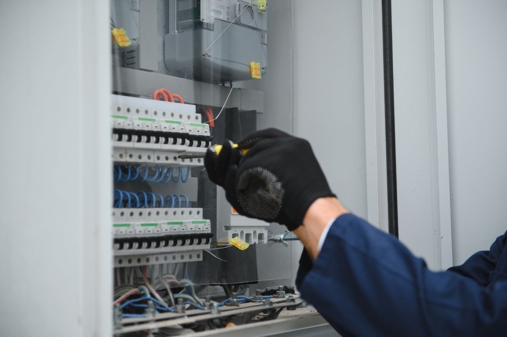 Electrician in a blue shirt and black gloves working on a fuse box.