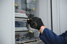 Electrician in a blue shirt and black gloves working on a fuse box.