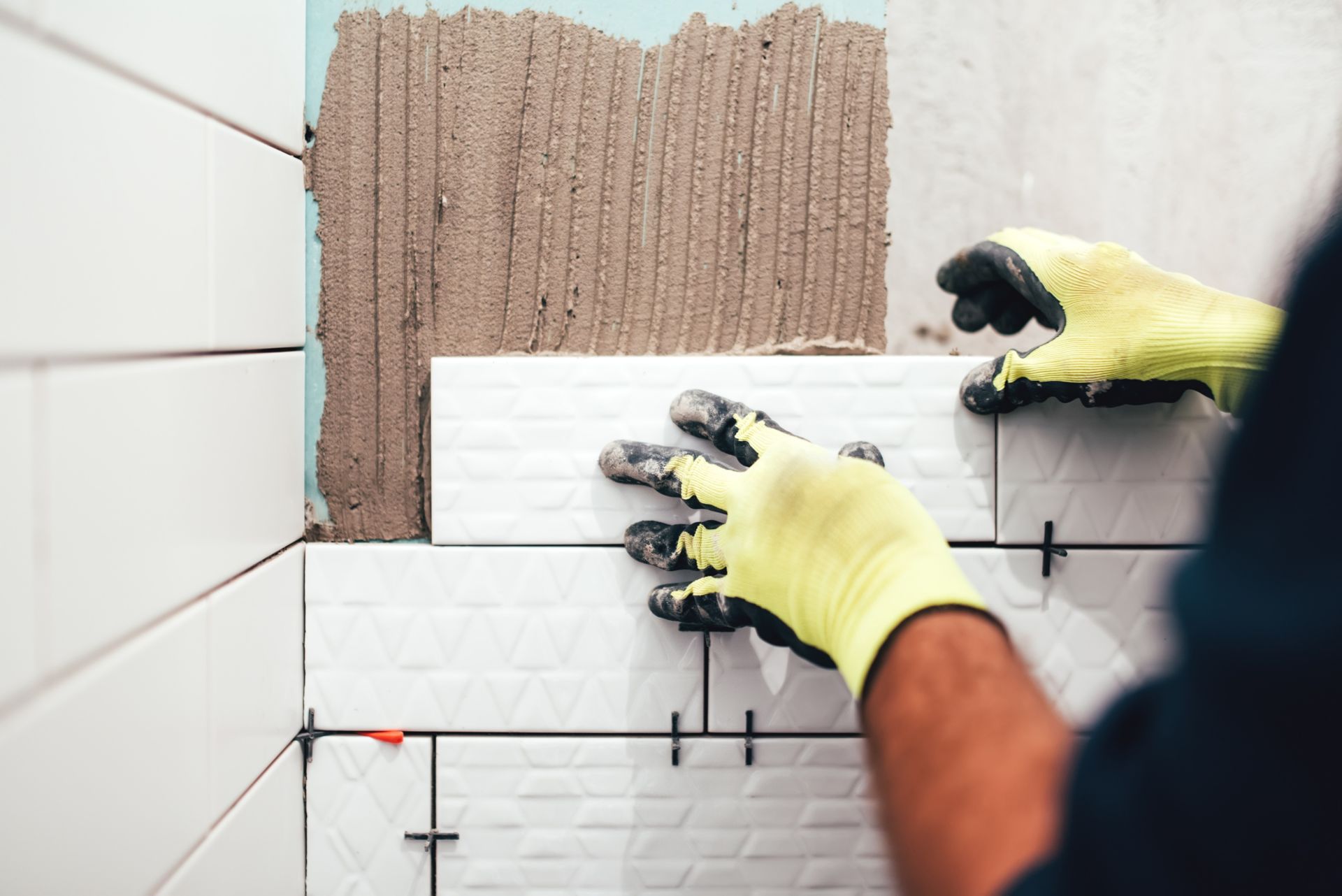 Gloved hands installing white wall tiles over brown adhesive in a tiled bathroom