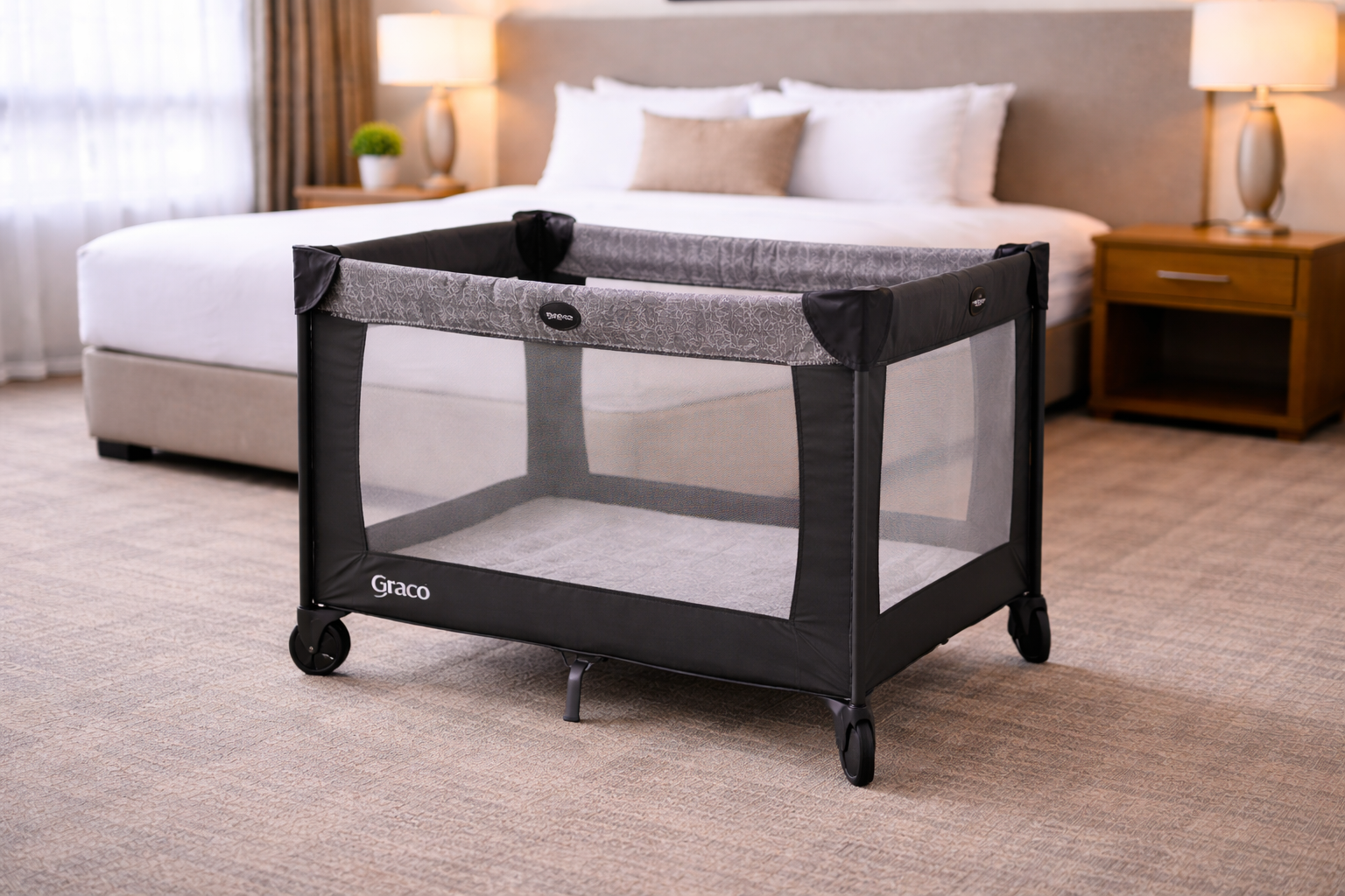 A portable gray and black playard standing on a carpeted floor in a bedroom with a bed and nightstands in the background.