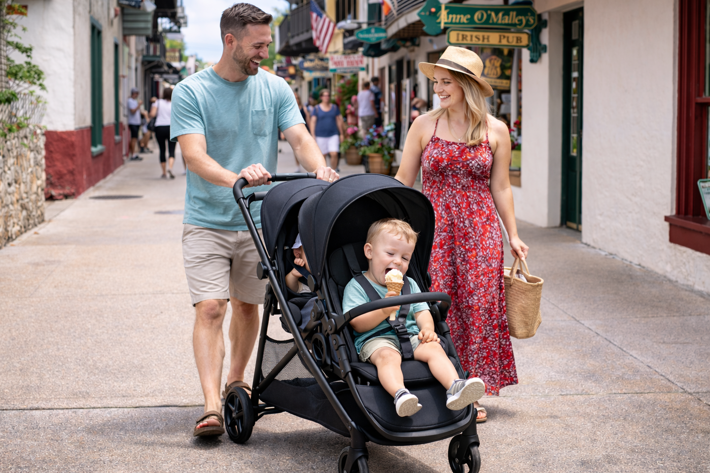 A family walks down a sunny outdoor street; a person pushes a double stroller while a child inside eats ice cream.