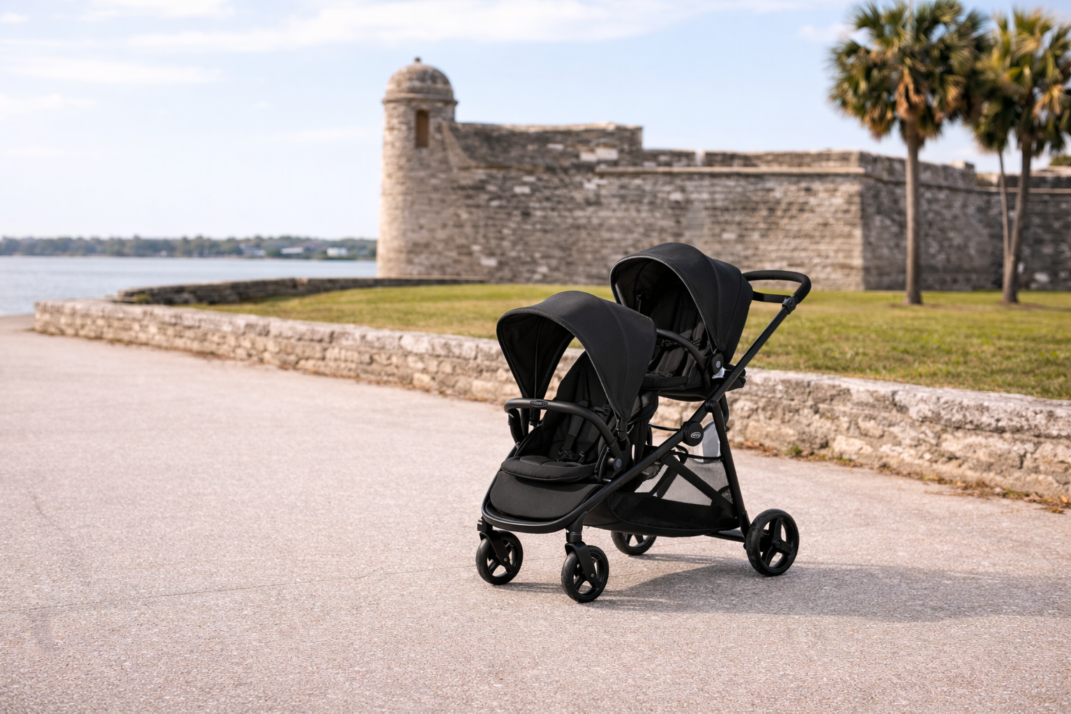 A black double stroller parked on a paved path in front of a historic stone fortress and palm trees by the water.