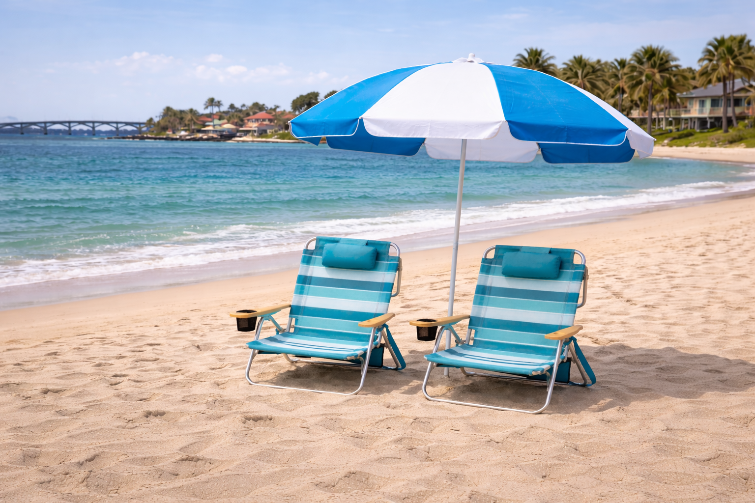 Two blue and white striped beach chairs sit under a matching beach umbrella on a sunny, sandy shoreline near the ocean.