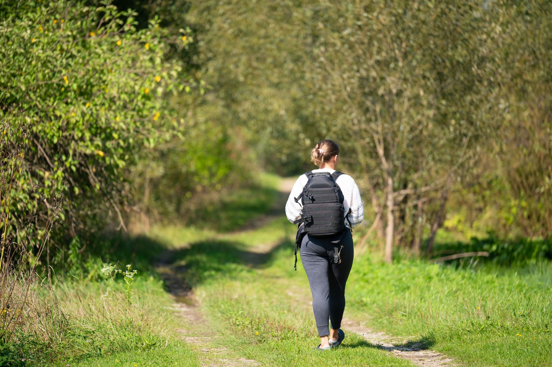 Woman hiking on a grassy trail, wearing a backpack, surrounded by trees and greenery.