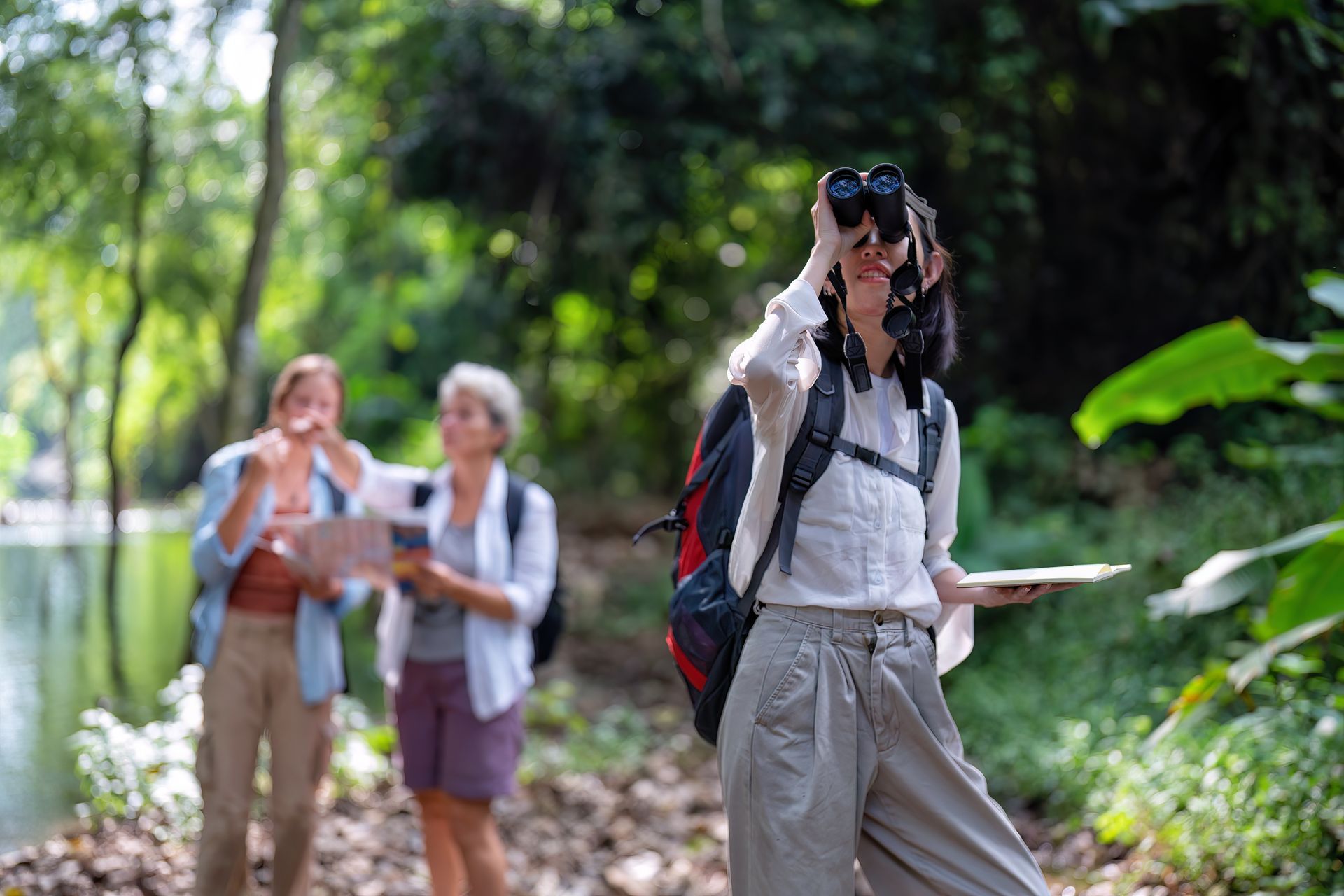 Woman with binoculars in a forest, holding a map, with two blurred figures behind her.