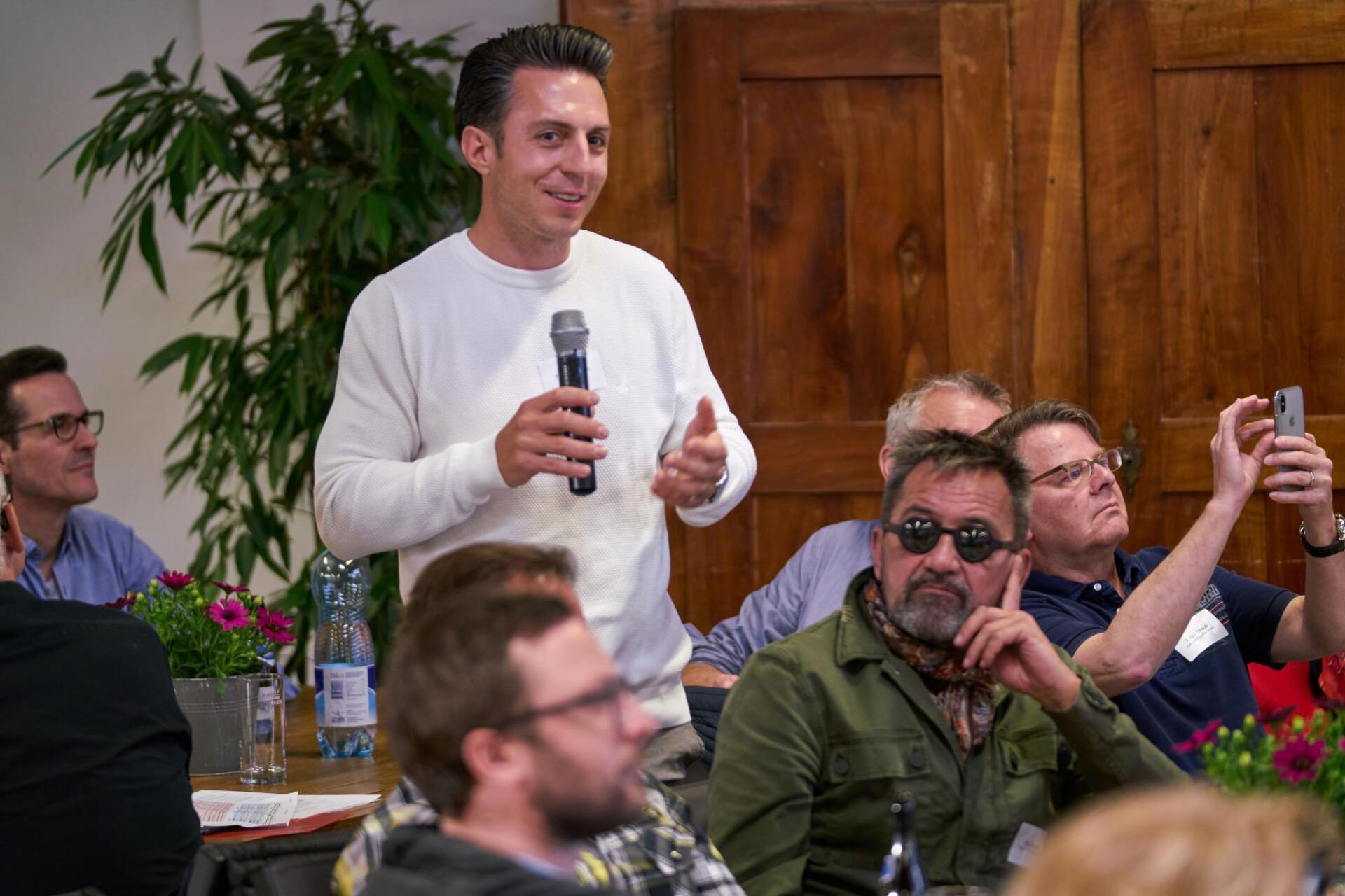 Ein Mann hält vor einer Gruppe von Menschen ein Mikrofon.