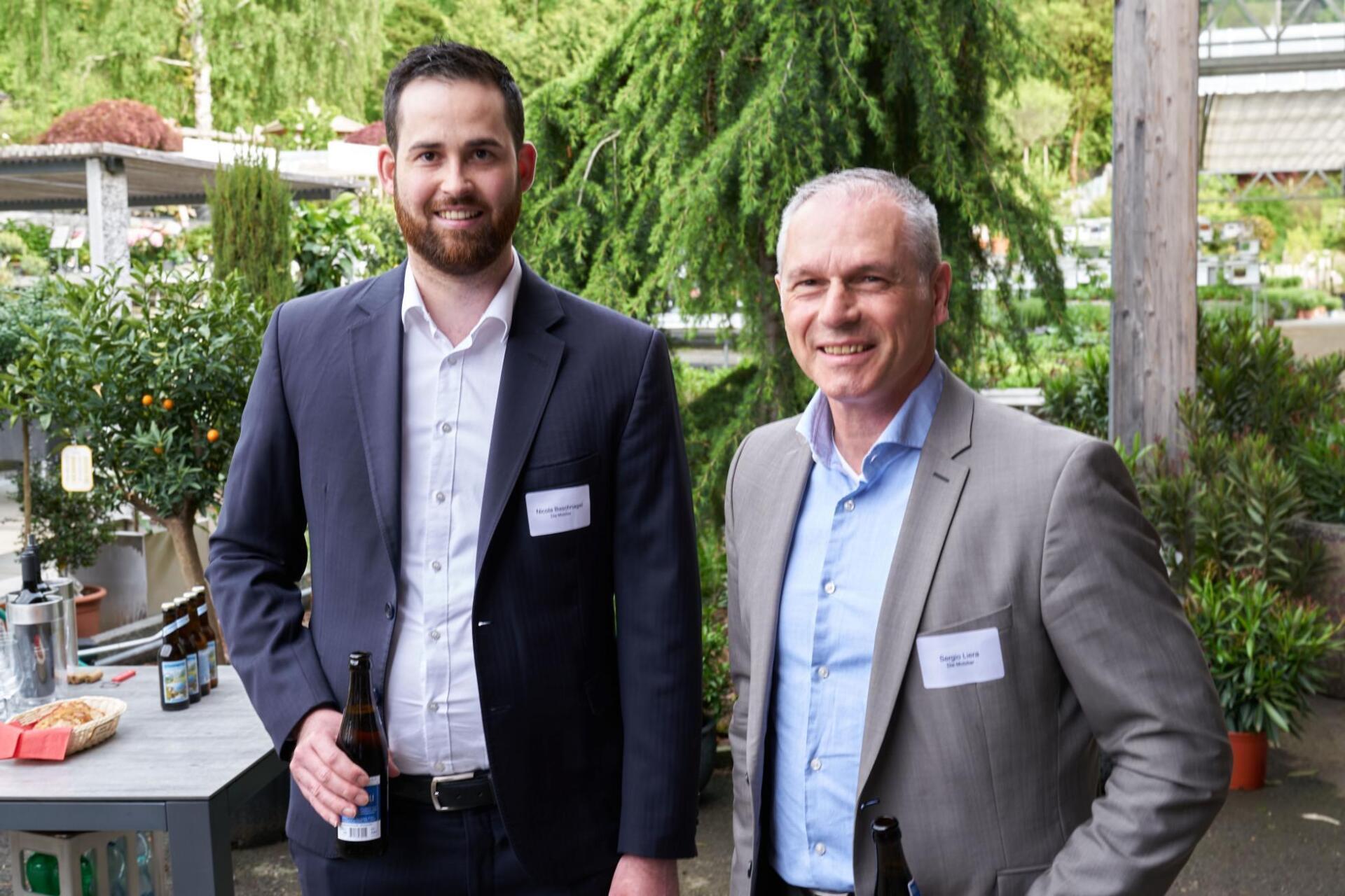 Zwei Männer in Anzügen stehen nebeneinander und halten Bierflaschen in den Händen.
