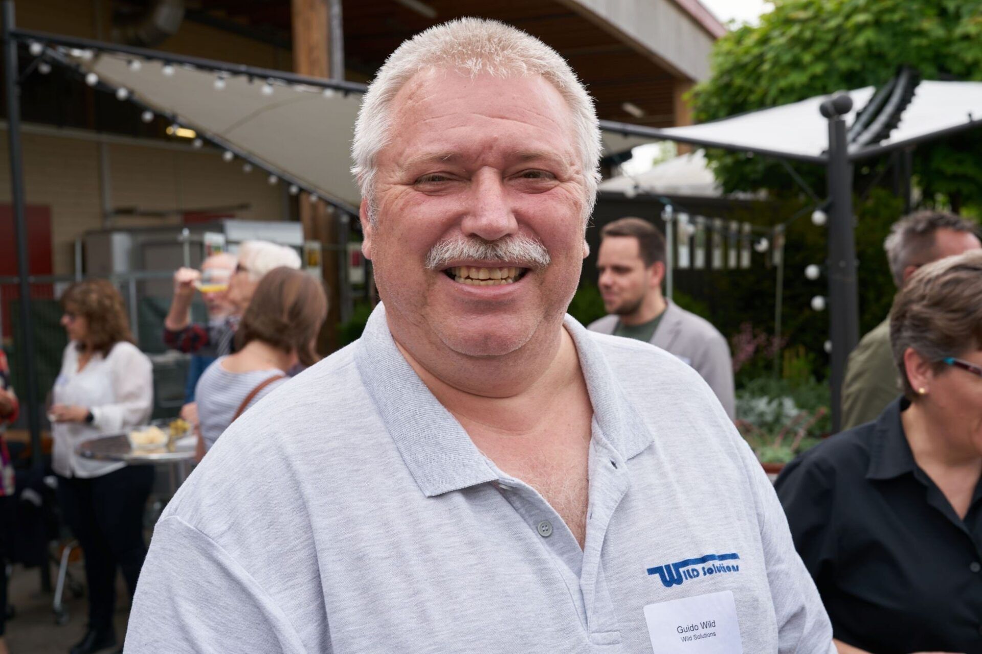 Ein Mann mit Schnurrbart lächelt vor einer Gruppe von Menschen.