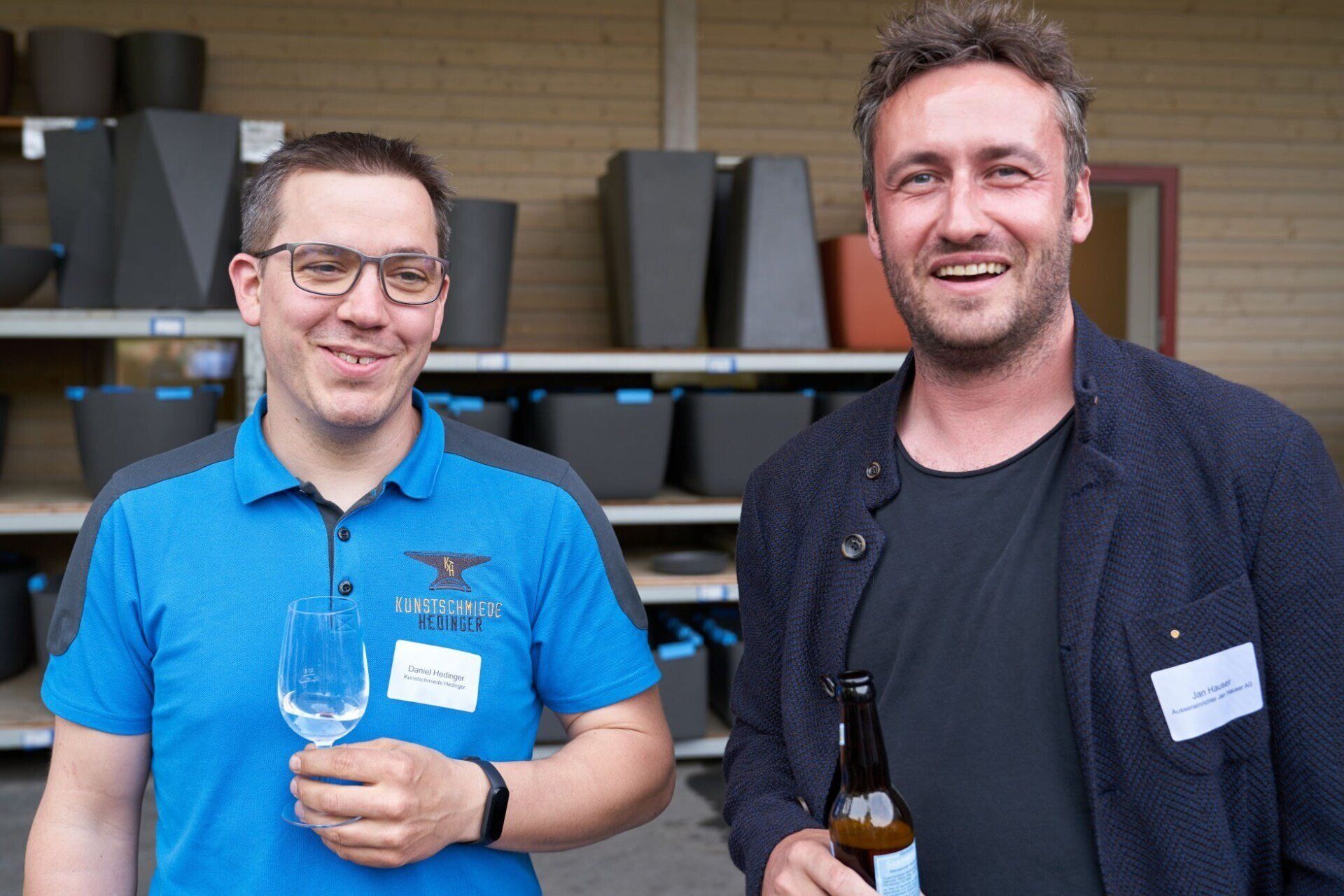 Zwei Männer stehen nebeneinander. Einer hält ein Weinglas und der andere eine Bierflasche.