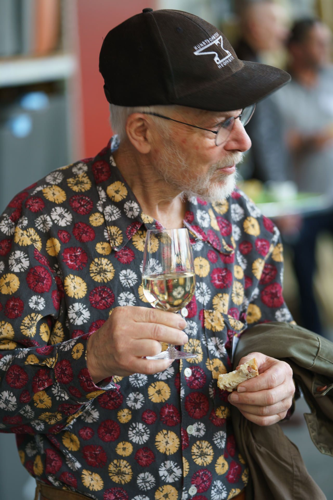 Ein Mann mit Hut und Brille hält ein Glas Wein.
