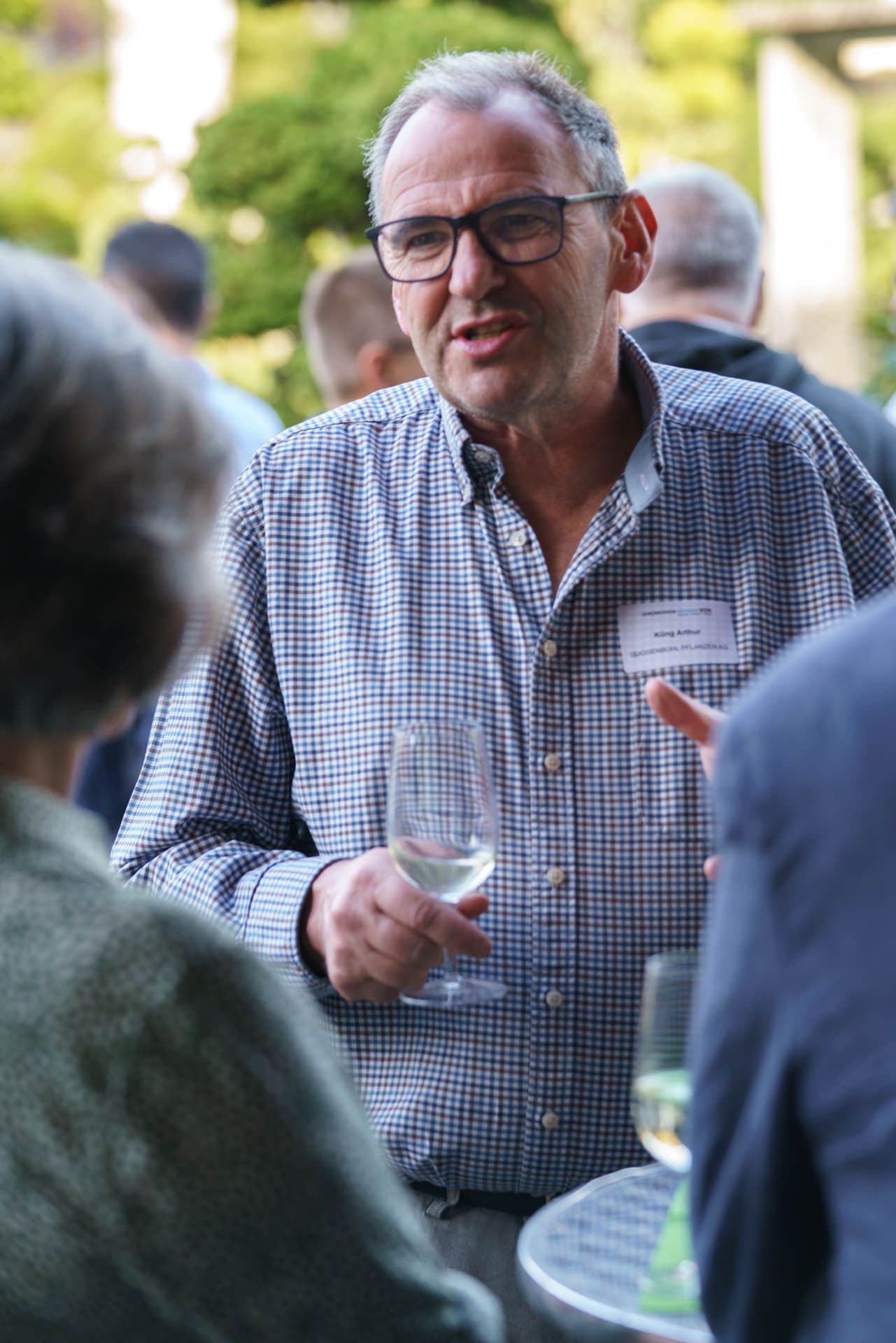 Ein Mann im karierten Hemd hält ein Glas Wein und unterhält sich mit einem anderen Mann.