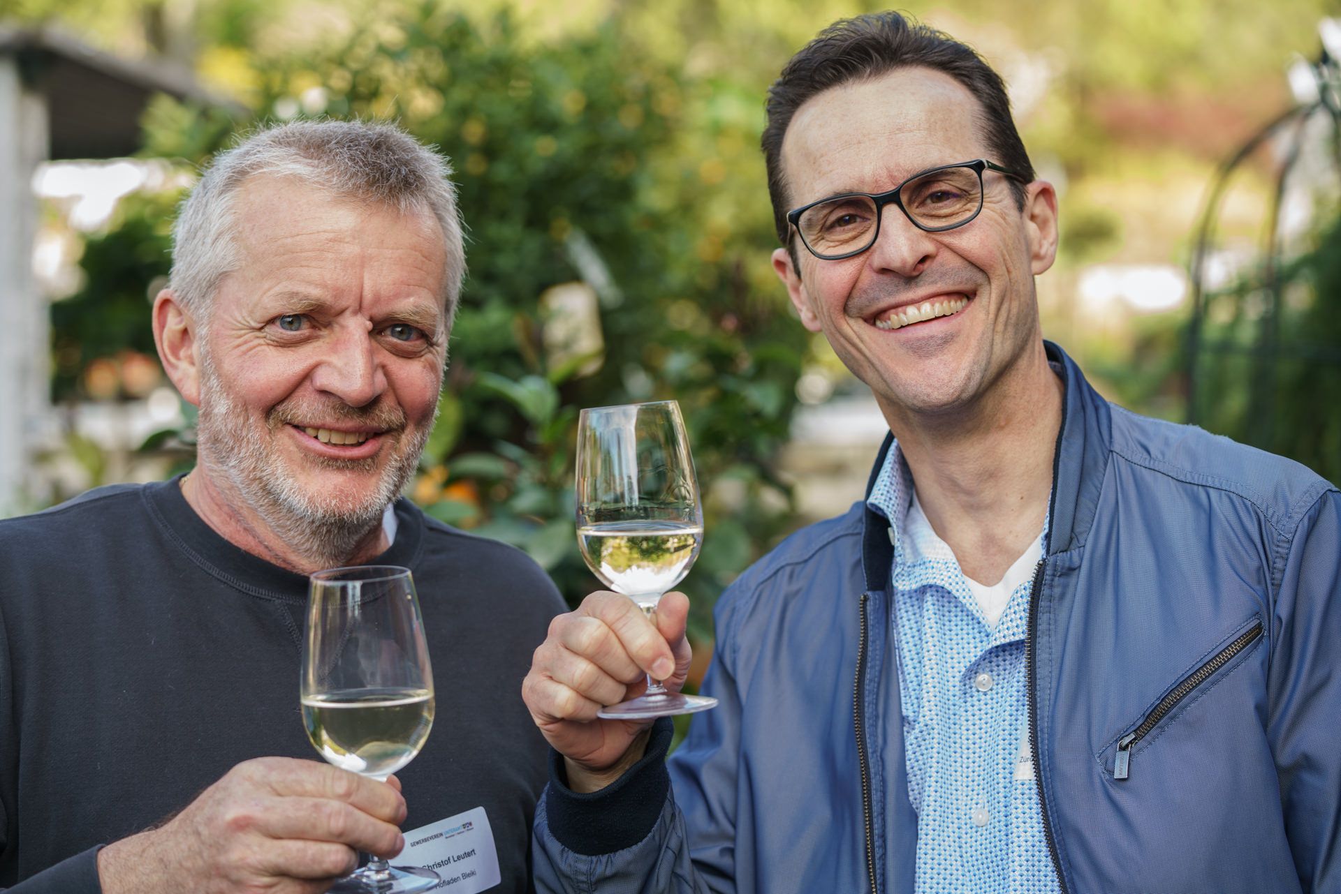 Zwei Männer halten Weingläser und lächeln in die Kamera.