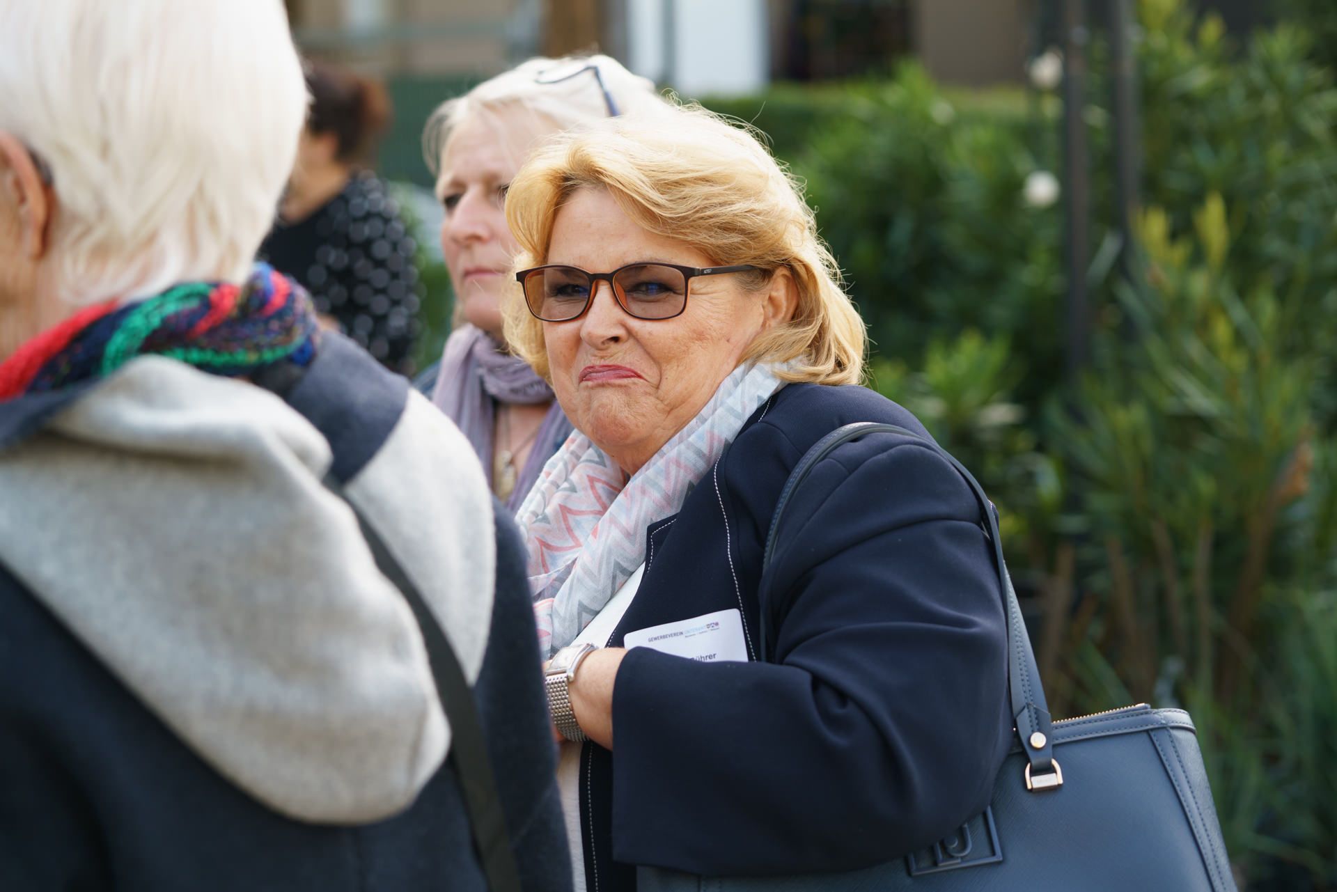 Eine Frau mit Brille und Handtasche unterhält sich mit einer anderen Frau.