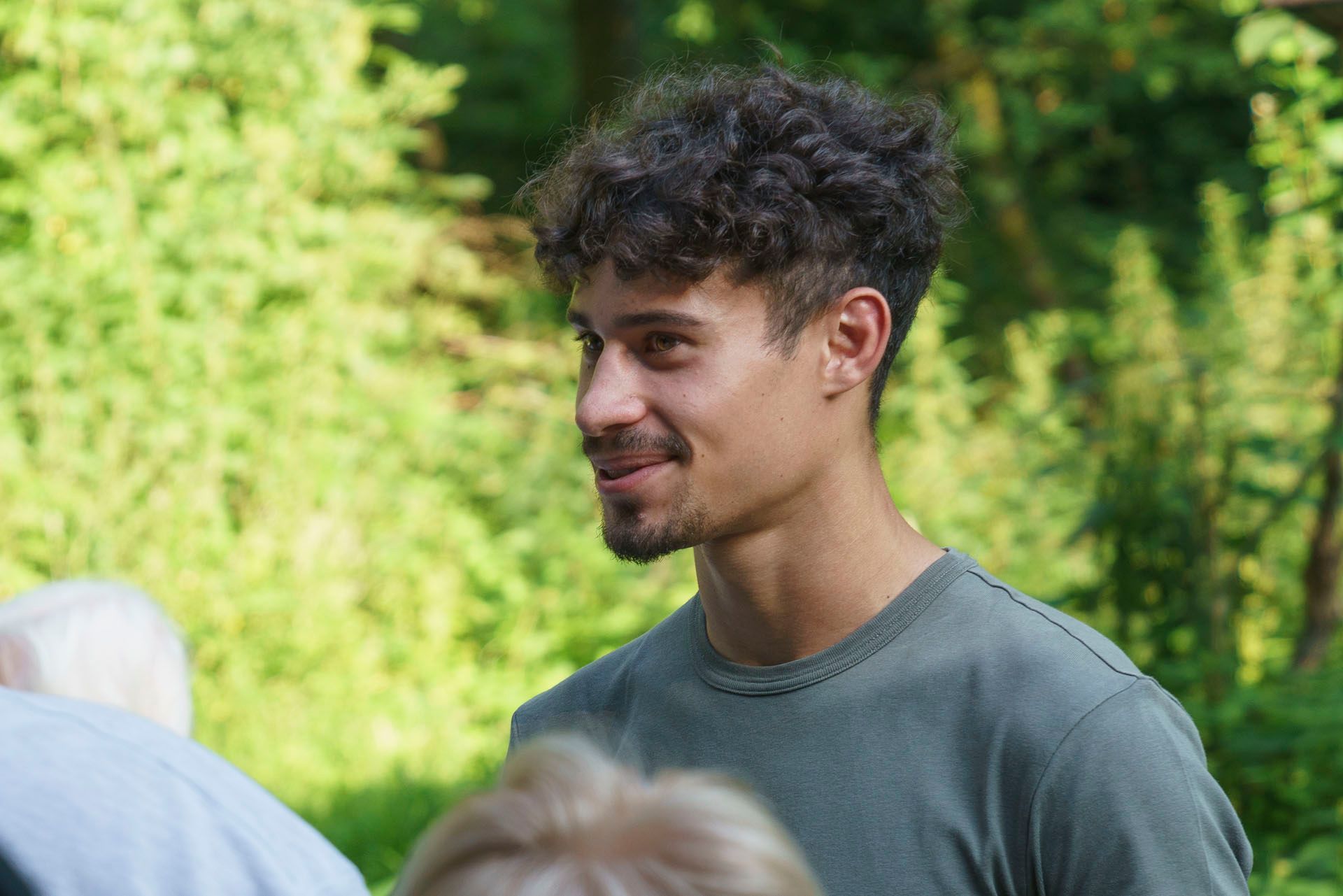 Ein Mann mit lockigem Haar und Bart steht vor einer Gruppe von Menschen.
