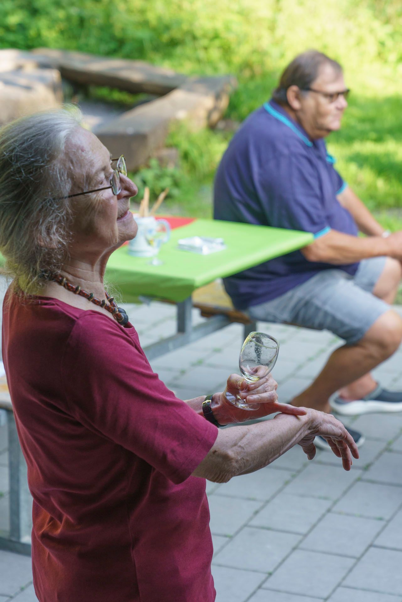 Eine ältere Frau hält ein Getränk, während ein Mann an einem Tisch sitzt.