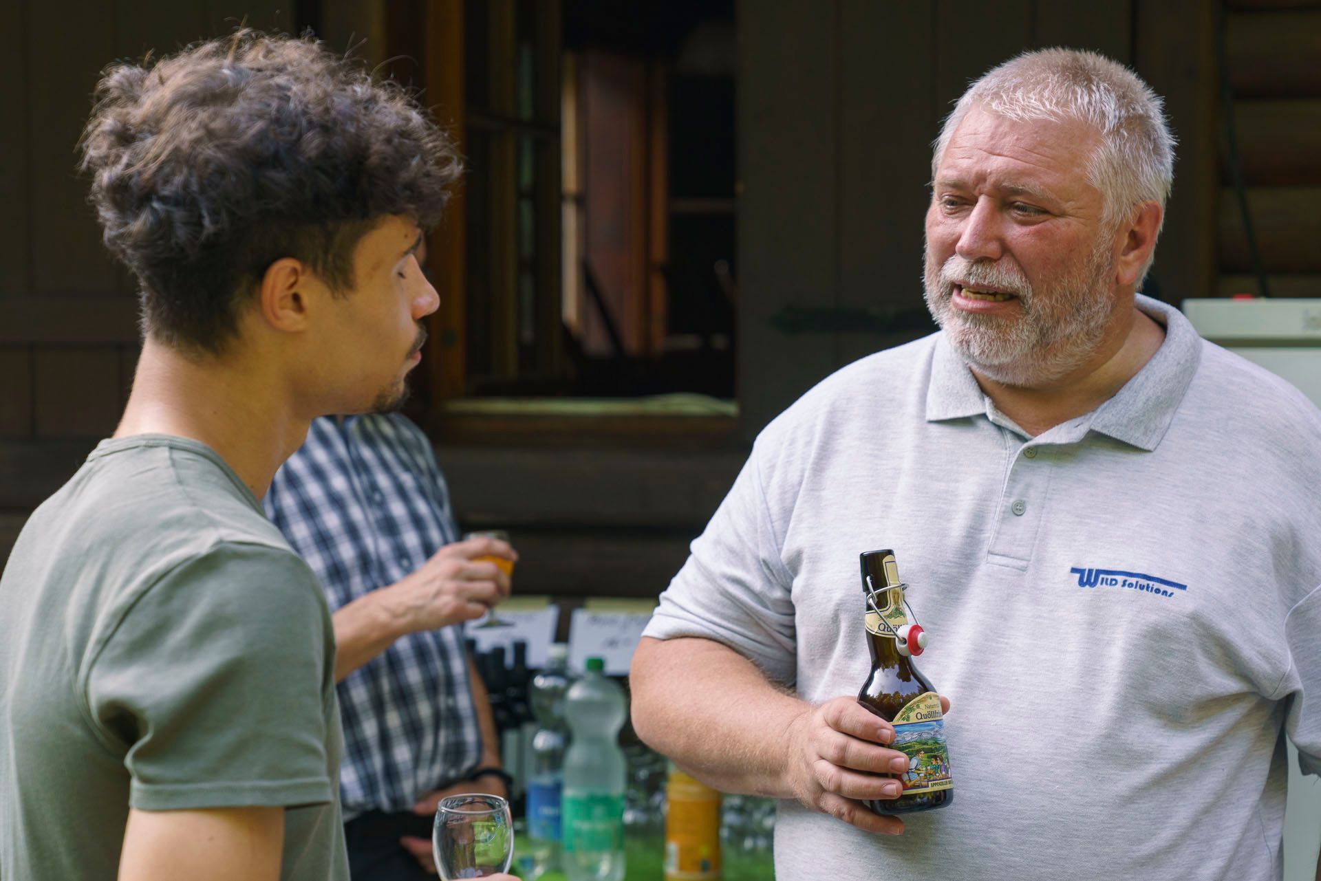 Ein Mann mit Bart hält eine Flasche Bier und spricht mit einem anderen Mann.