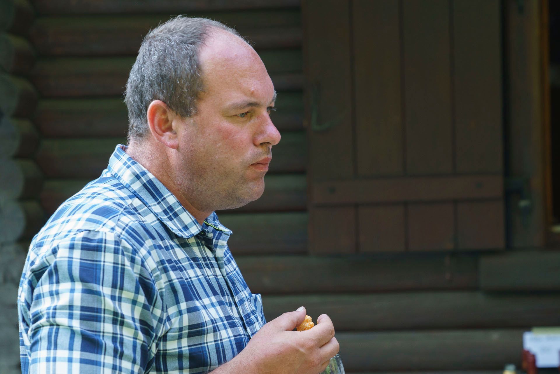 Ein Mann im karierten Hemd isst vor einer Blockhütte ein Sandwich.