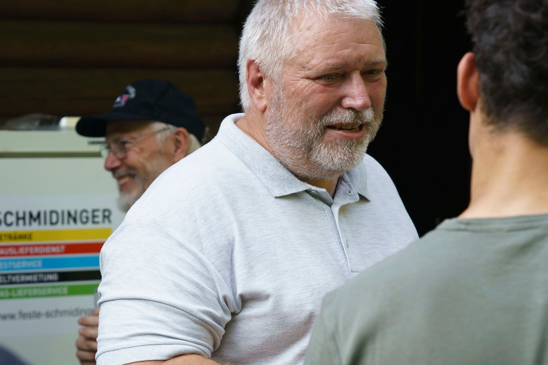 Ein Mann mit Bart spricht mit einem anderen Mann vor einem Schild mit der Aufschrift „Schmidtinger“