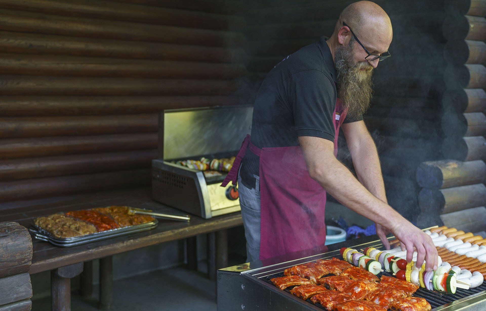 Ein Mann mit Bart bereitet Essen auf einem Grill vor einer Blockhütte zu.