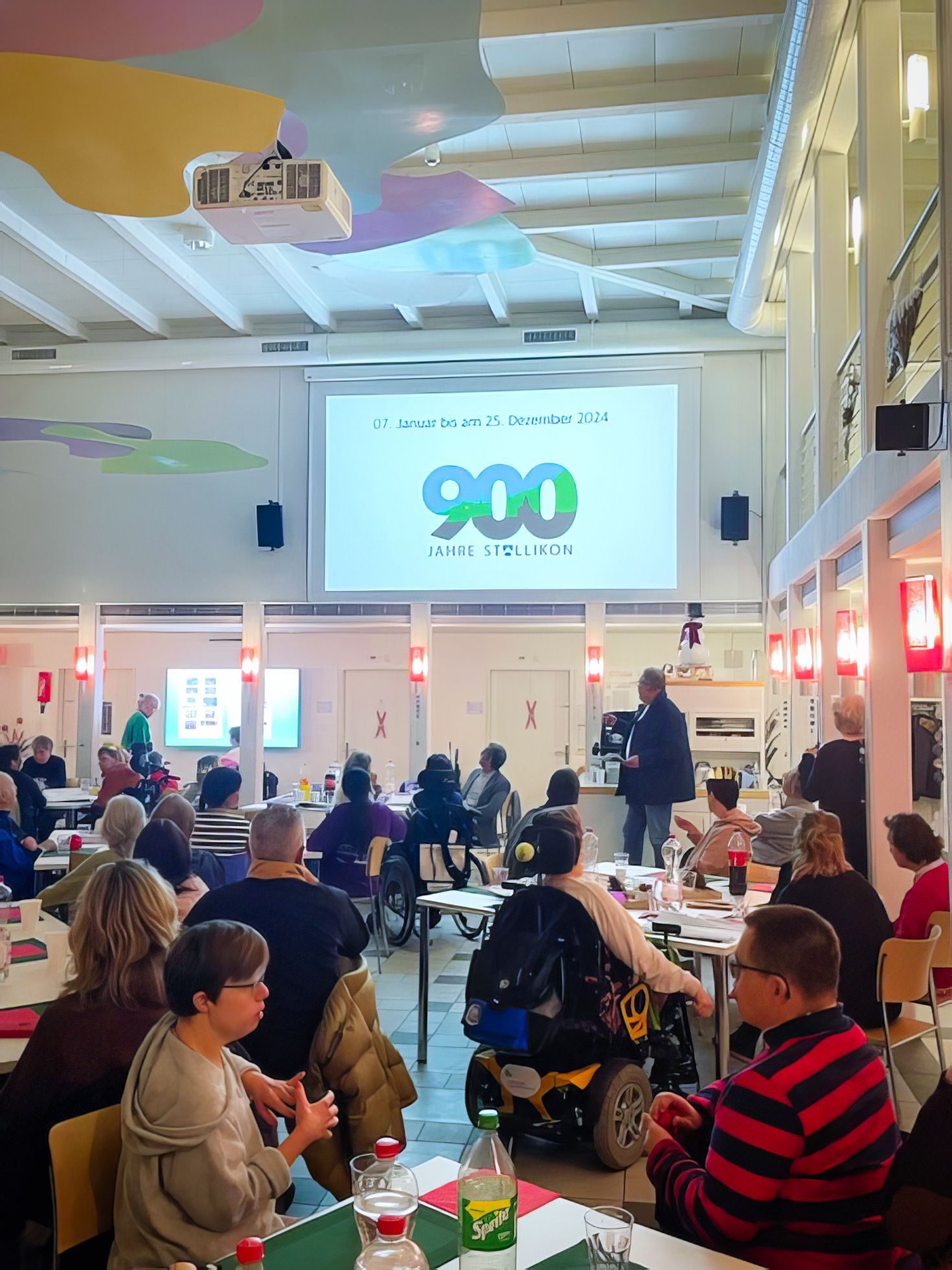 Eine Gruppe von Menschen sitzt an Tischen vor einem Bildschirm mit der Aufschrift 900