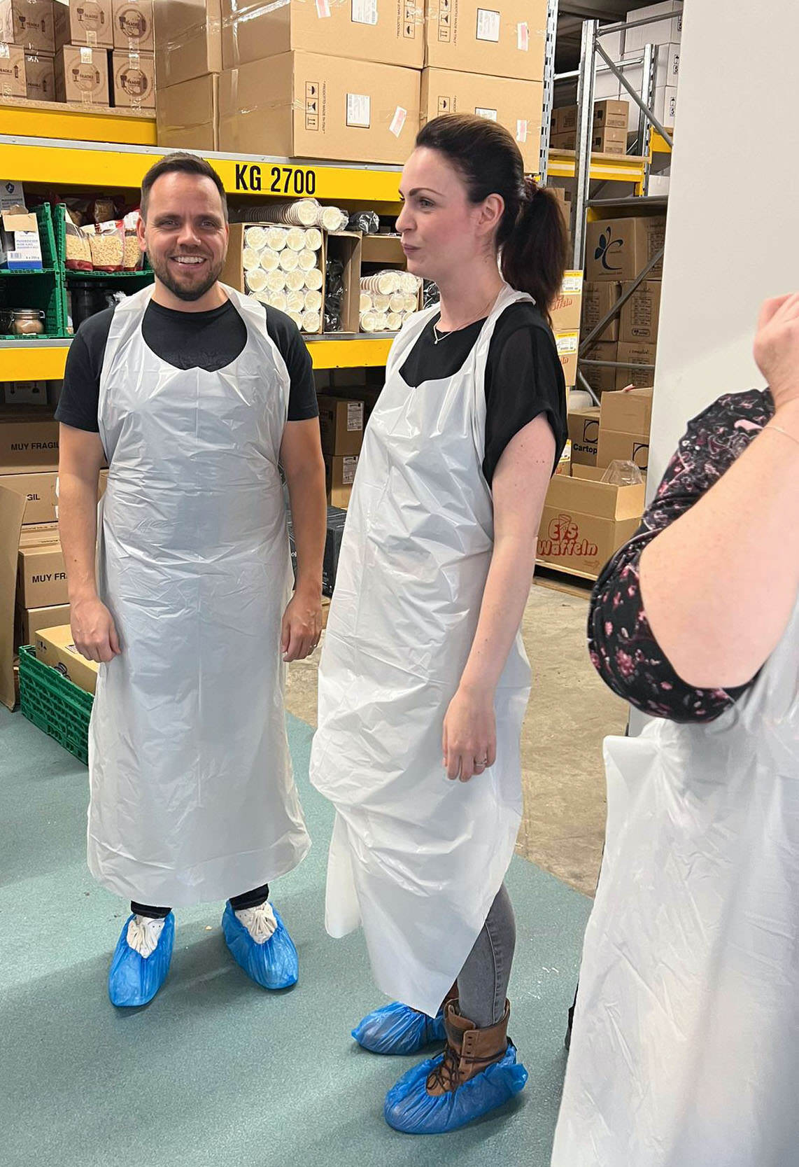 Ein Mann und eine Frau mit Schürzen und blauen Schuhüberzügen stehen in einer Lagerhalle.