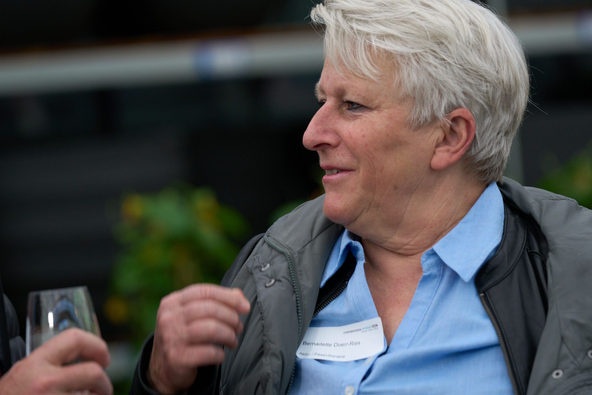 Ein Mann mit weißem Haar hält ein Glas Wein und unterhält sich mit einem anderen Mann.