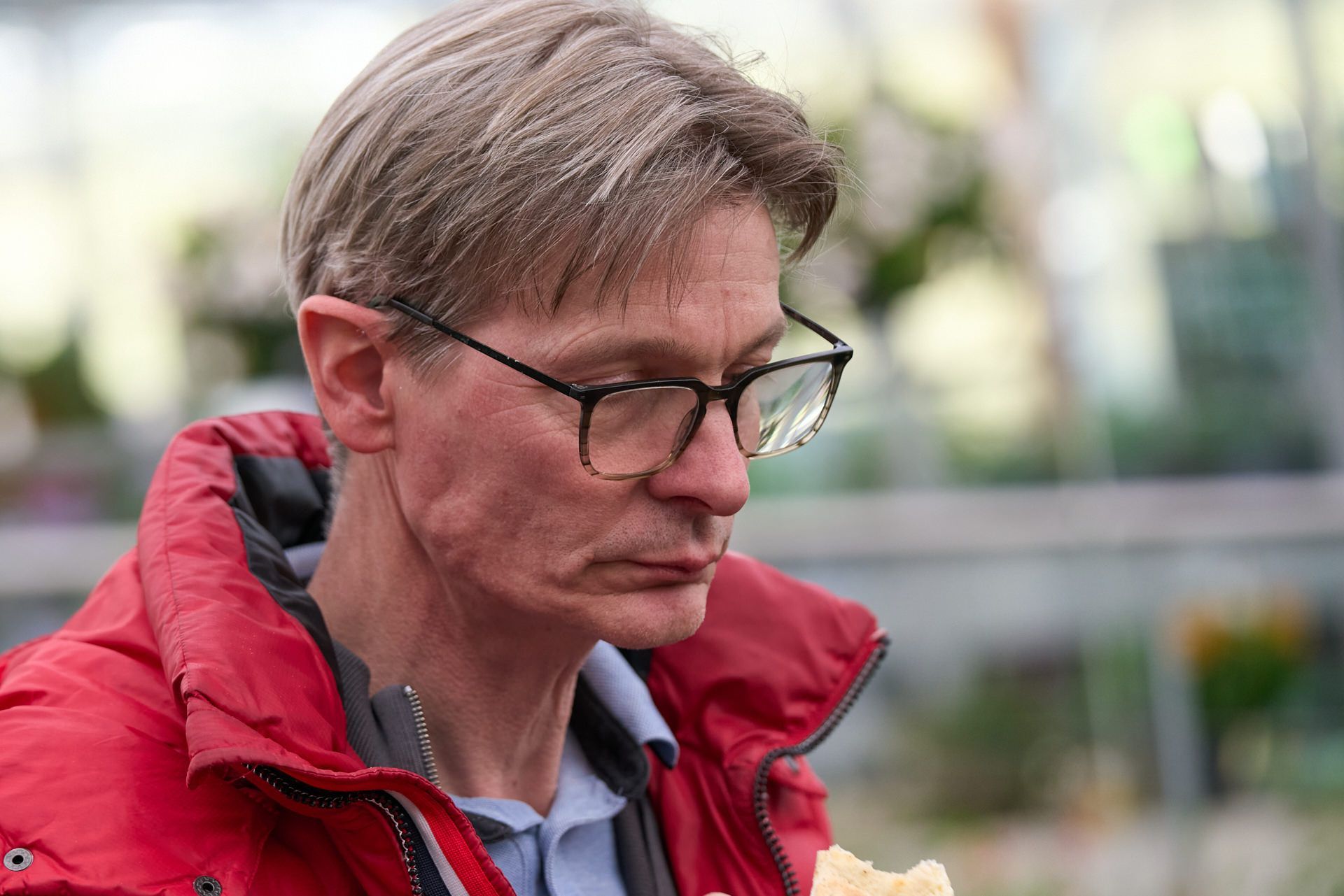 Ein Mann mit Brille und roter Jacke isst ein Sandwich.