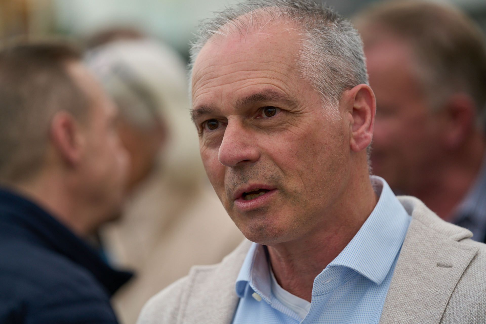 Ein Mann in einem blauen Hemd und einer hellbraunen Jacke spricht in einer Menschenmenge mit einem anderen Mann.