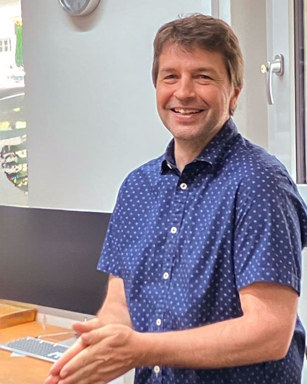 Ein Mann in einem blauen Hemd steht vor einem Fenster und lächelt.