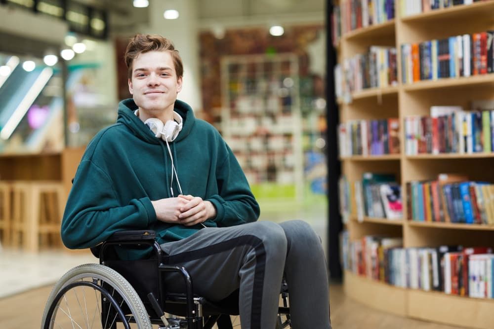 A Young Man is Sitting in a Wheelchair in a Library A Woman in a Wheelchair is Being Helped by a Man — Empowering You Disability Services In Maryborough, QLD