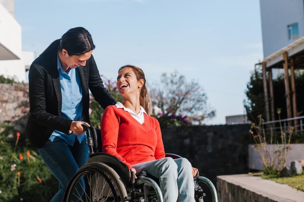 A Woman in a Wheelchair is Laughing While a Man Pushes Her — Empowering You Disability Services In Maryborough, QLD

