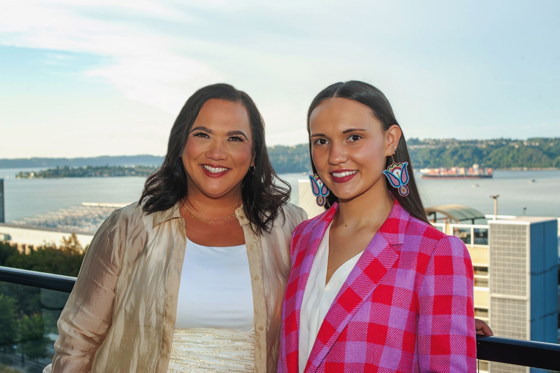 Two women smiling, on a balcony overlooking water. One in pink plaid blazer, the other in gold shirt.