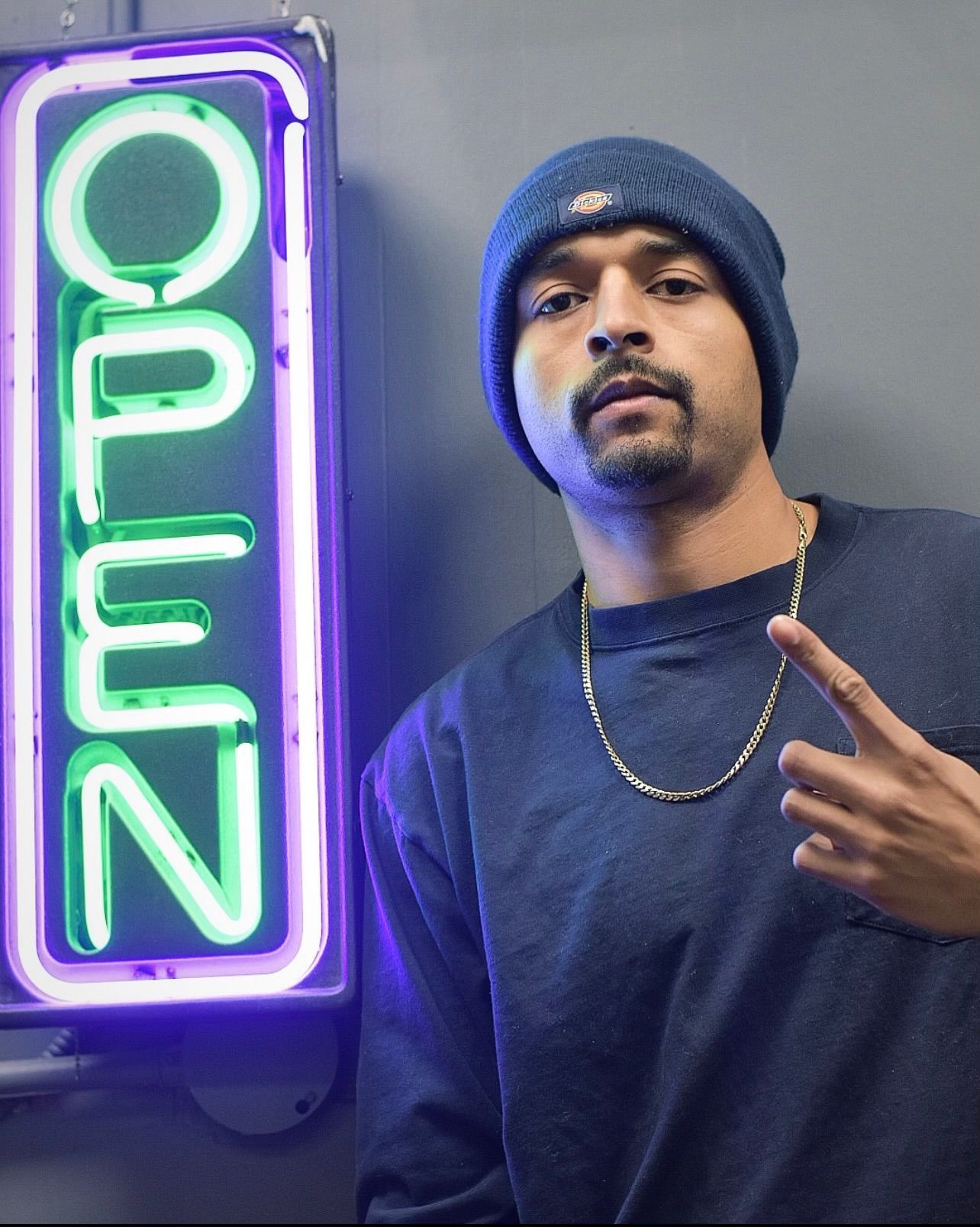 Man in beanie and sweatshirt stands by a neon
