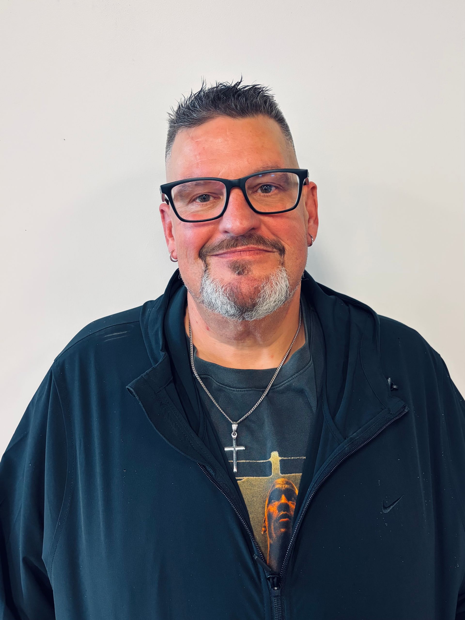 Man with glasses and goatee wearing a dark jacket and a cross necklace.