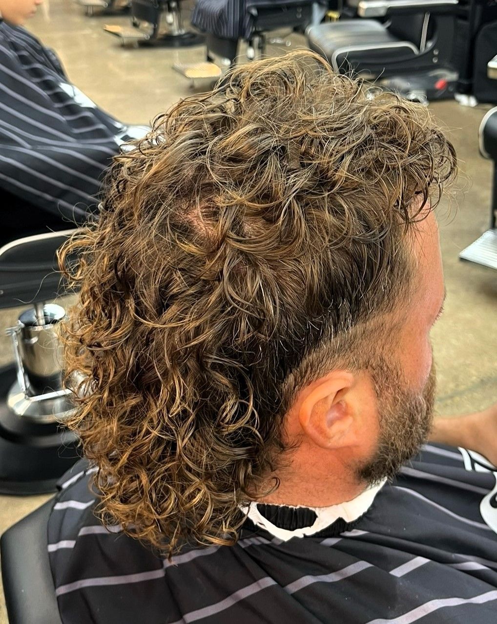 Man with curly, light brown hair styled in a mullet at a barbershop. Side profile.