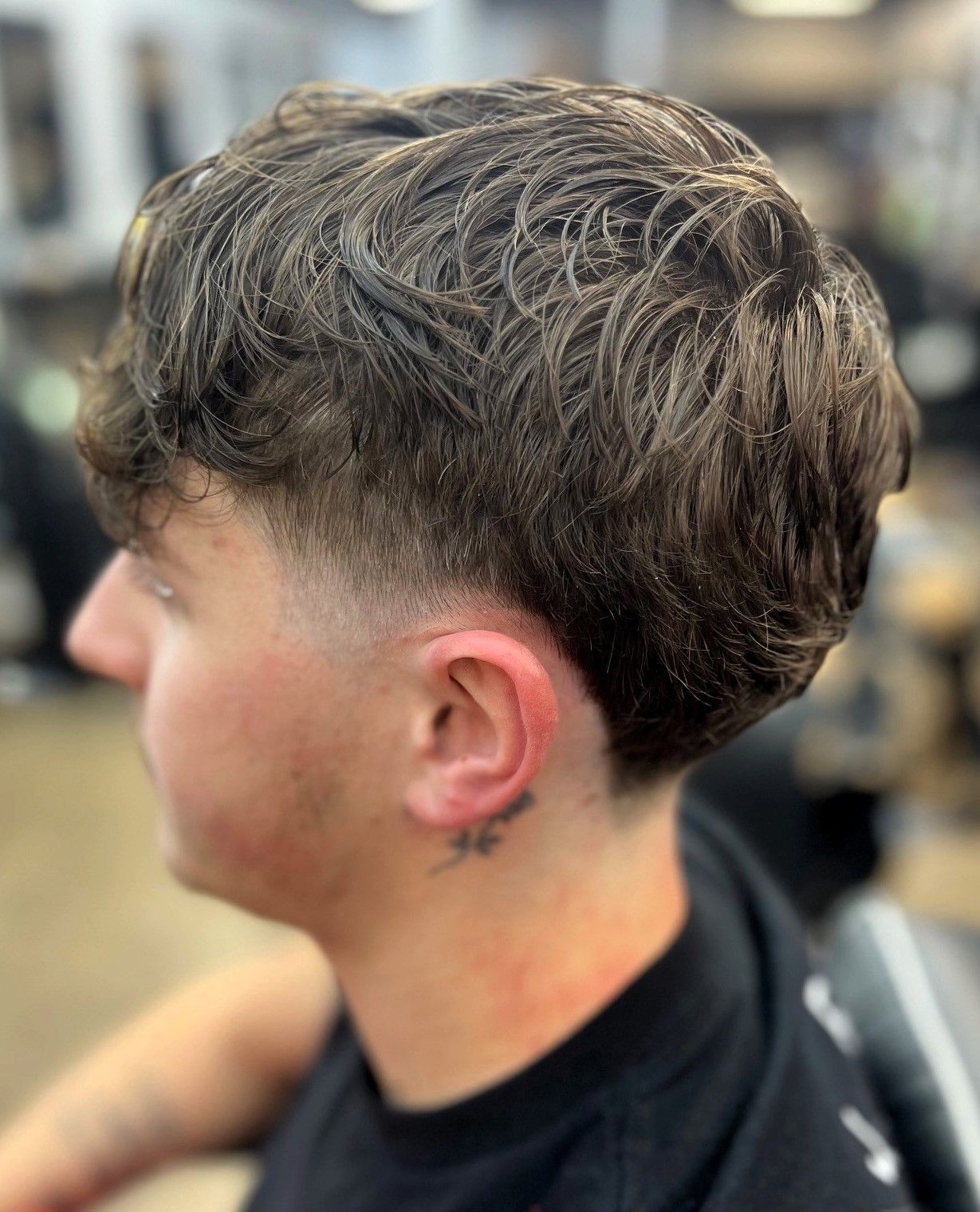 Man with a textured hairstyle and faded sides, viewed in profile. He has a neck tattoo.